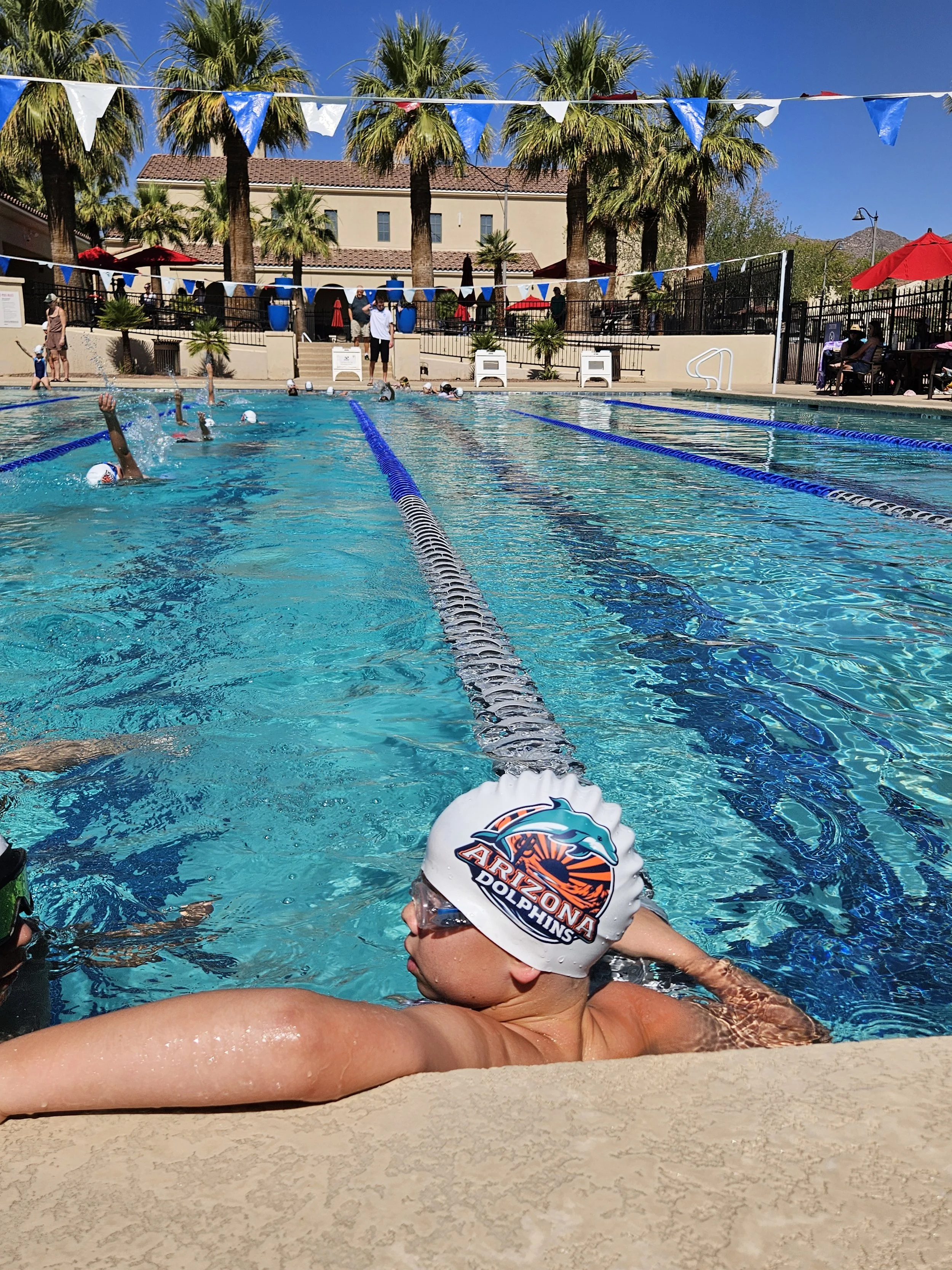 Swimmer with a white cap with the Arizona Dolphins logo, at an outdoor pool, preparing for a swim race.