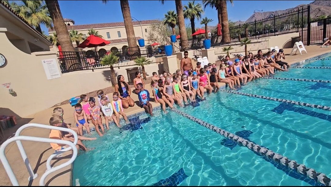 Group of children sitting on the edge of an outdoor swimming pool with a swimming instructor standing nearby, surrounded by palm trees and umbrellas.
