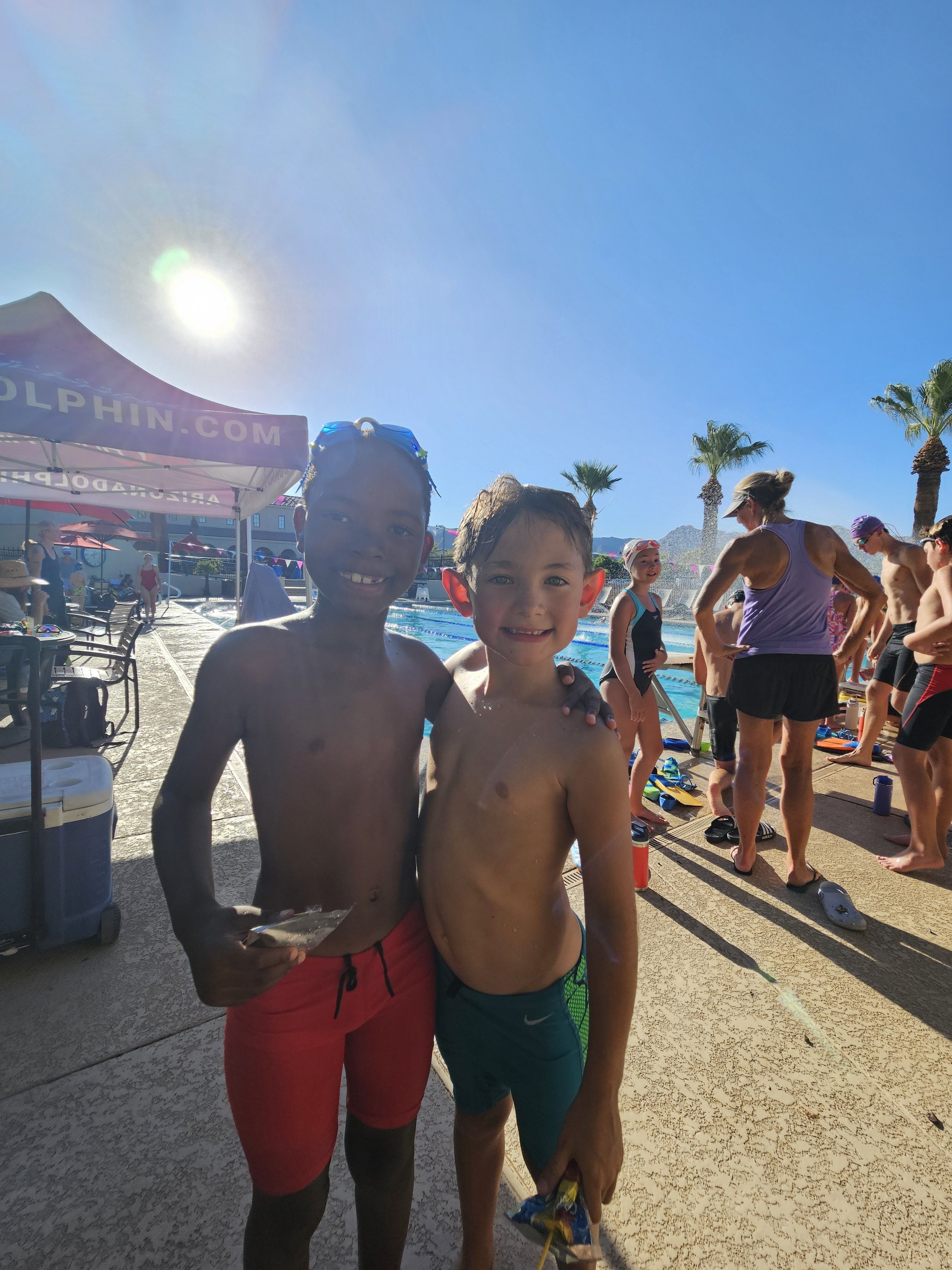 Two young boys in swim trunks standing by a pool with a crowd of people and palm trees in the background, during a sunny day.