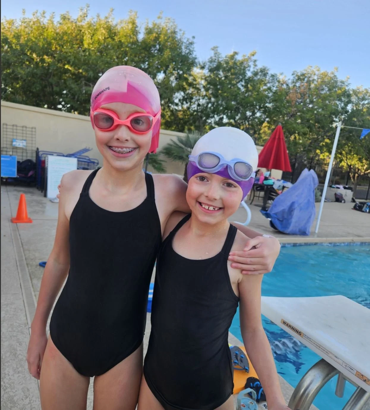 Two young girls in black swimsuits and swimming caps with goggles, smiling at the camera, at a swimming pool.