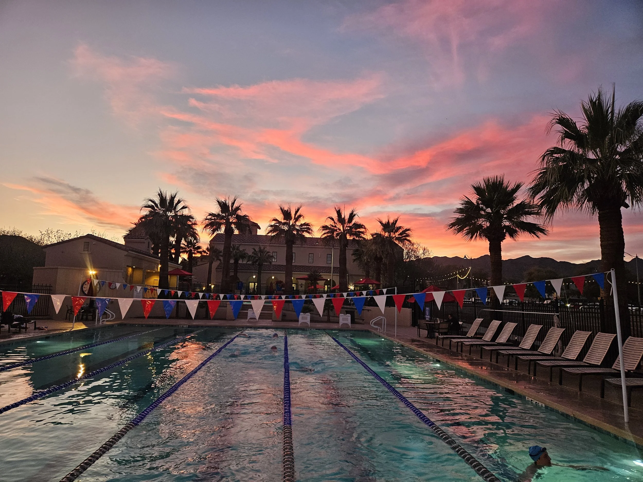 An outdoor swimming pool at sunset with decorative flags along the fence, surrounded by lounge chairs, palm trees, and nearby buildings with string lights. The sky is pink and purple with clouds.