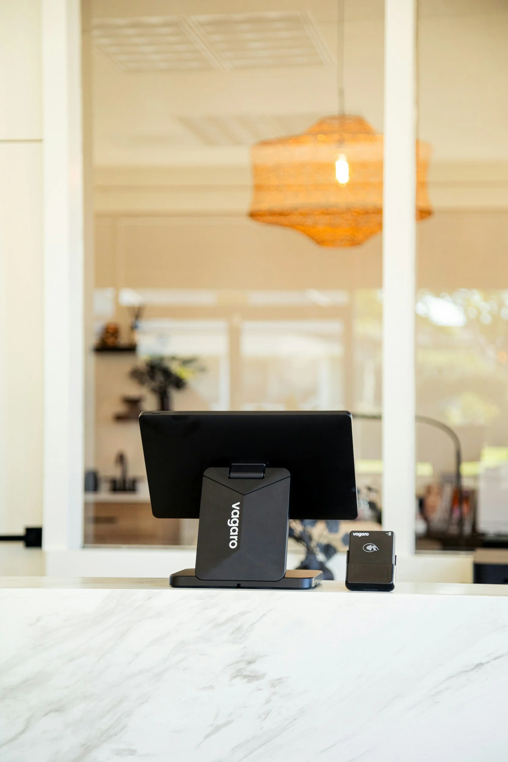 A modern office desk setup featuring a black Vigaro monitor and a Vagaroo wireless charging device on a white marble surface, with a blurred background of a home interior and warm hanging ceiling lights.
