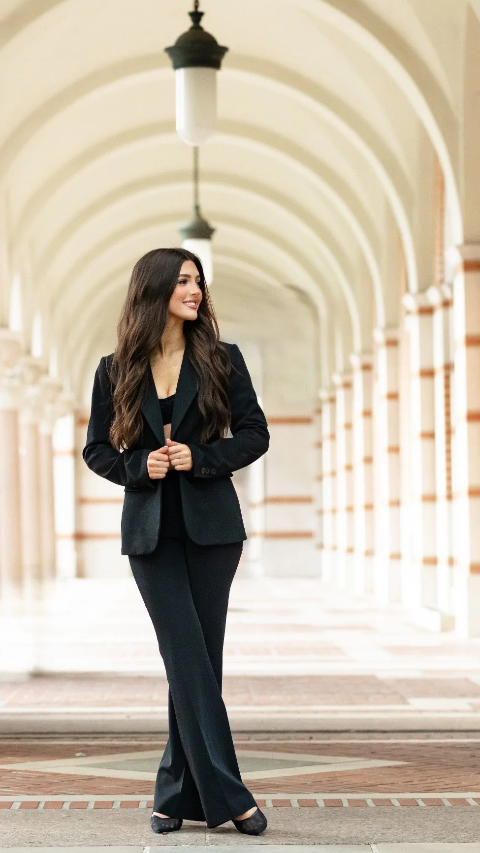 A woman in a black pantsuit standing in an arched corridor with hanging lights.