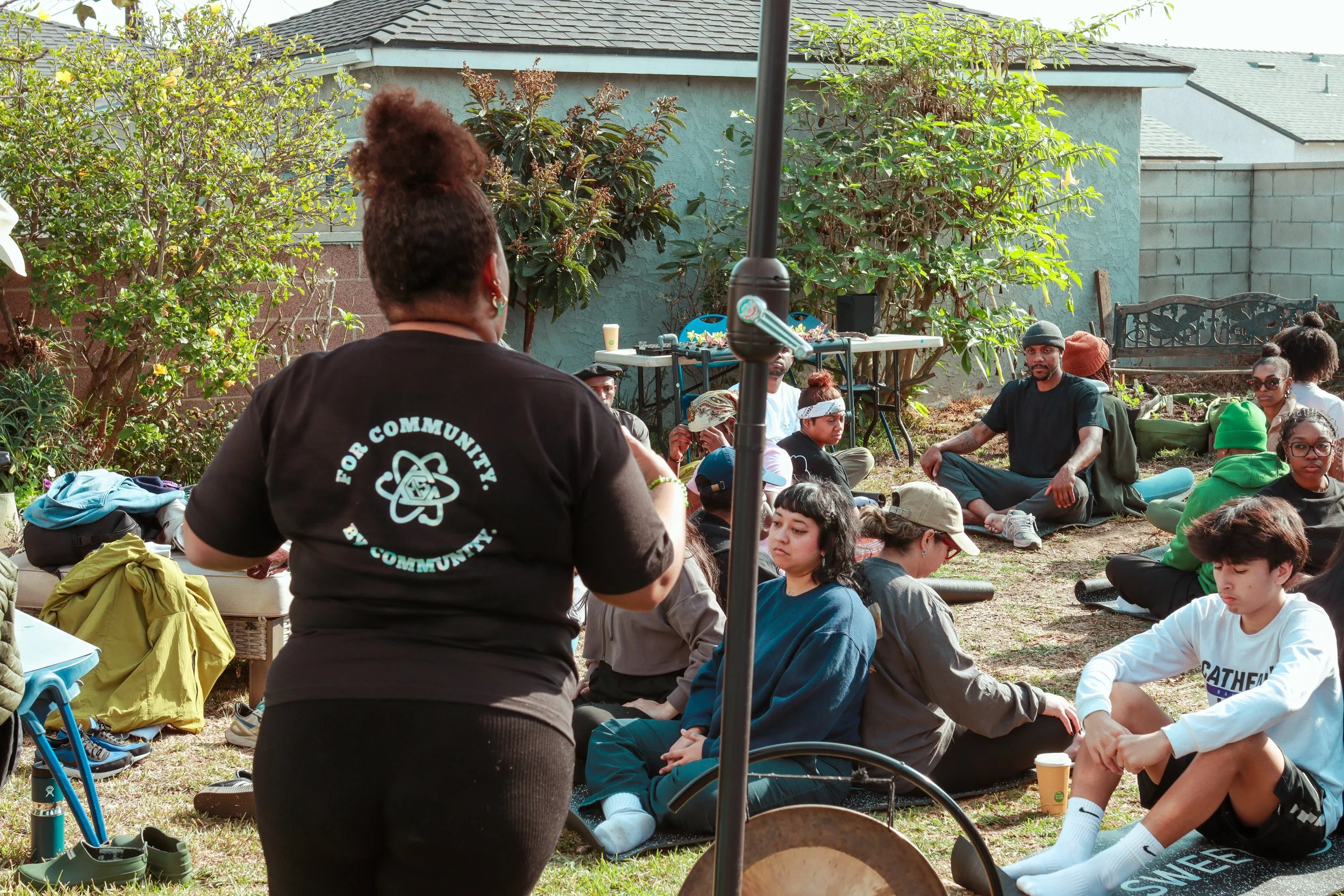 A group of people sitting on the grass in a backyard, attending a community gathering or event, with some seated in meditation or listening to a speaker. A woman in black stands in the foreground, facing the seated audience.