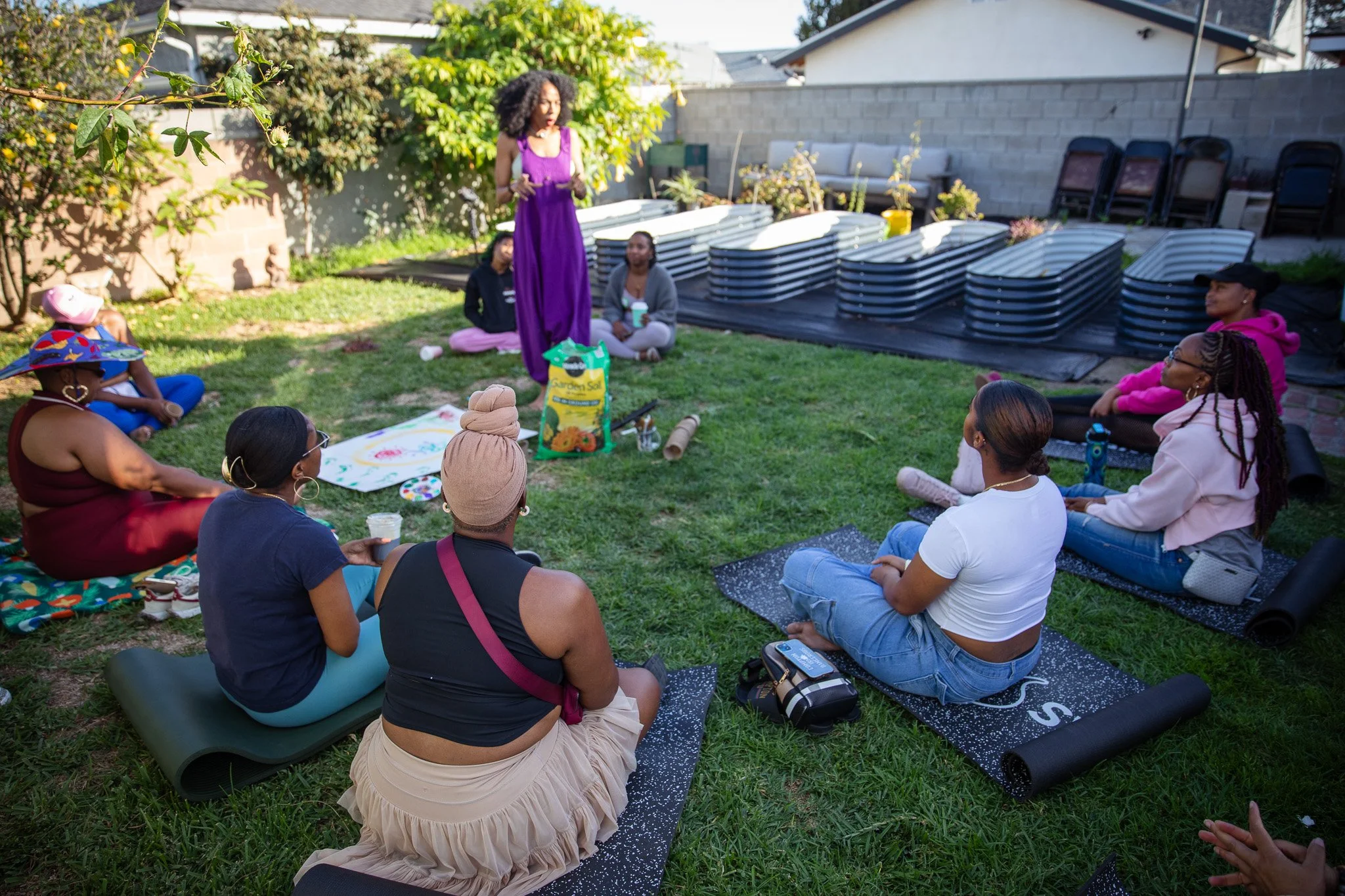 A group of women sitting on yoga mats in a backyard, listening to a woman in purple speaking, with gardening supplies and stacked patio furniture behind them.