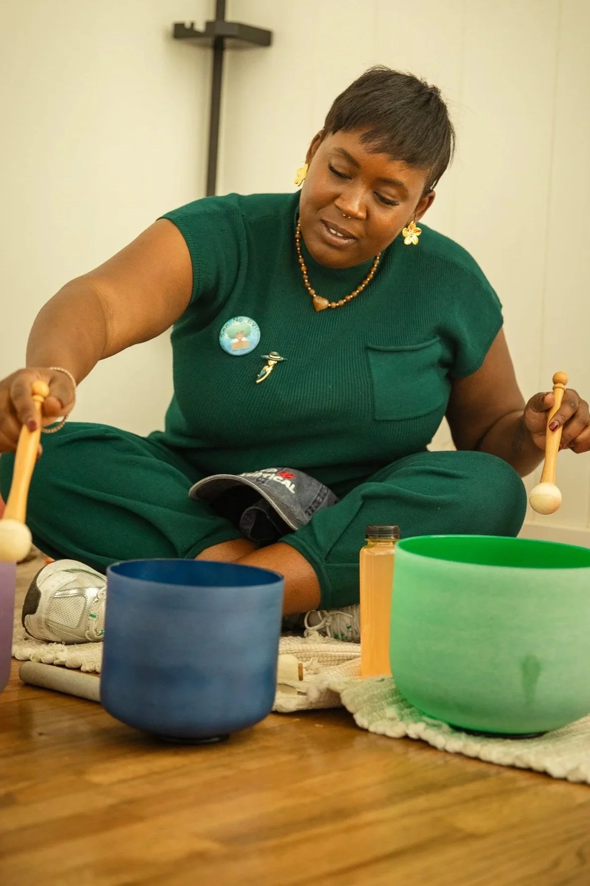 A woman sitting cross-legged on the floor playing a hand drum with colorful bowls and a small bottle nearby.