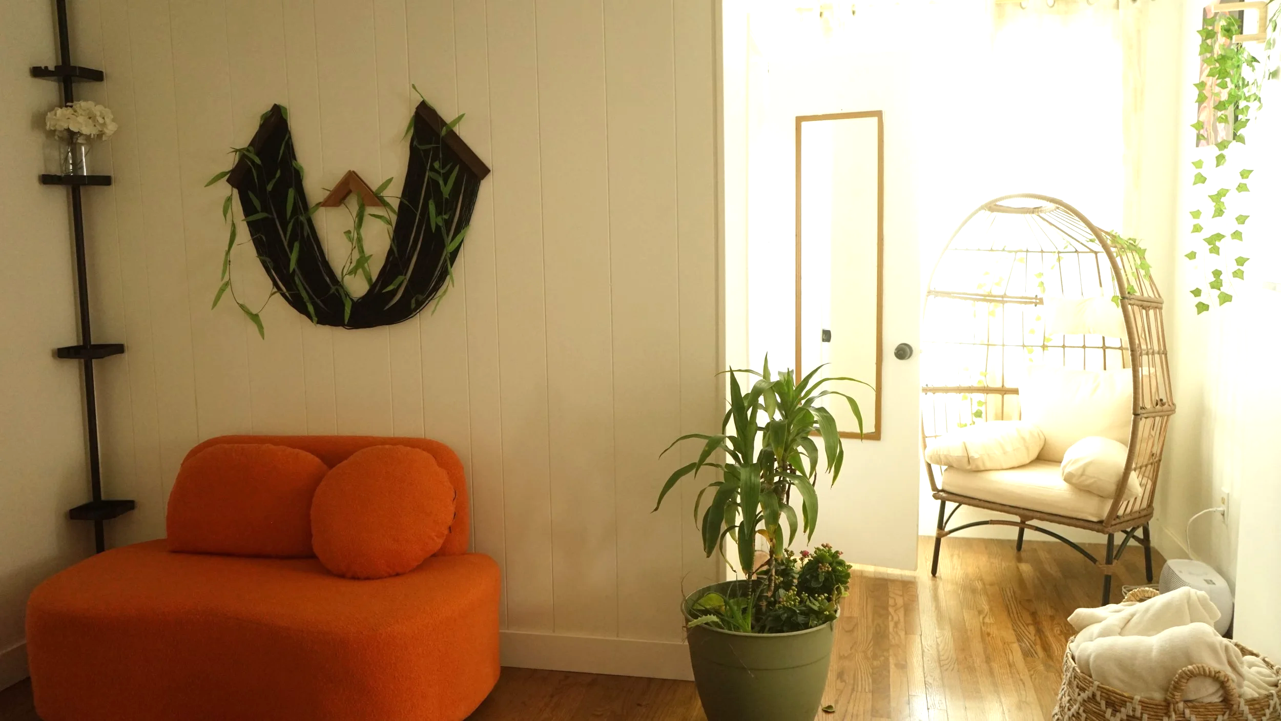 Cozy indoor space with an orange loveseat, a potted green plant, a hanging egg chair with cushions near a doorway, and decorative wall art.