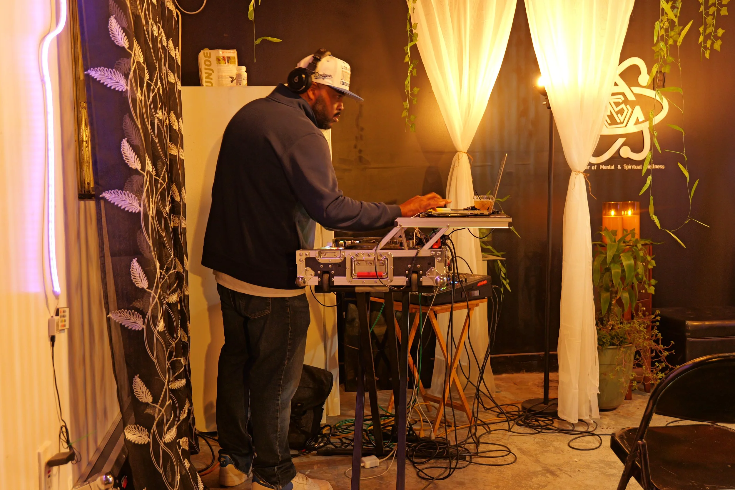DJ mixing and using a laptop in a cozy, dimly lit room decorated with plants, candles, and curtains.