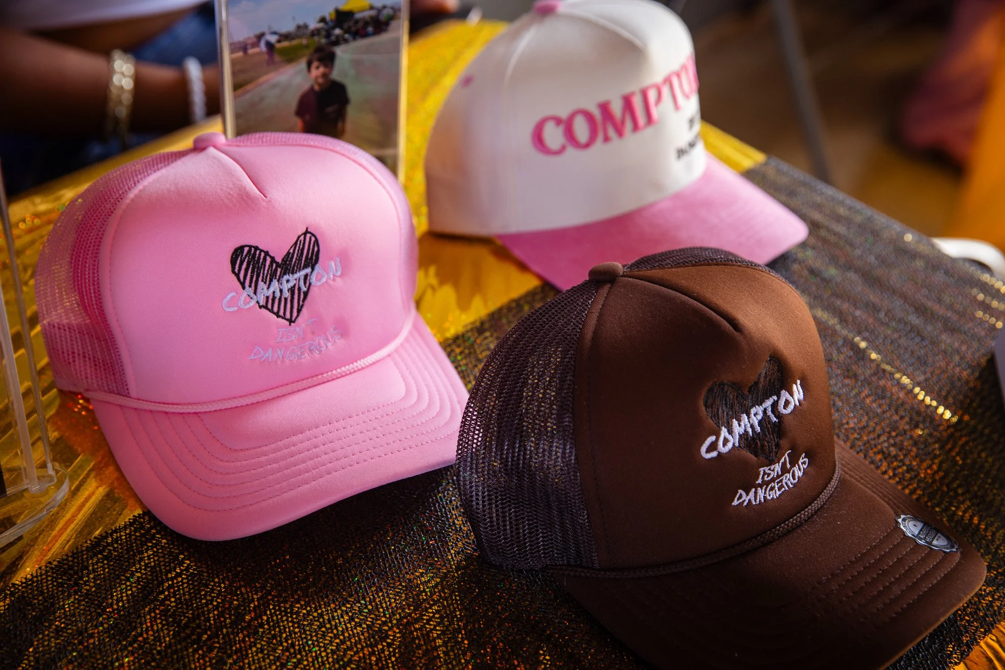 Three baseball caps on a table, each with embroidered text and designs, one pink with a heart and the words "COMPTON IS MY DANGEROUS," a white and pink with, "COMPTON," and a brown with the same design as the pink cap.