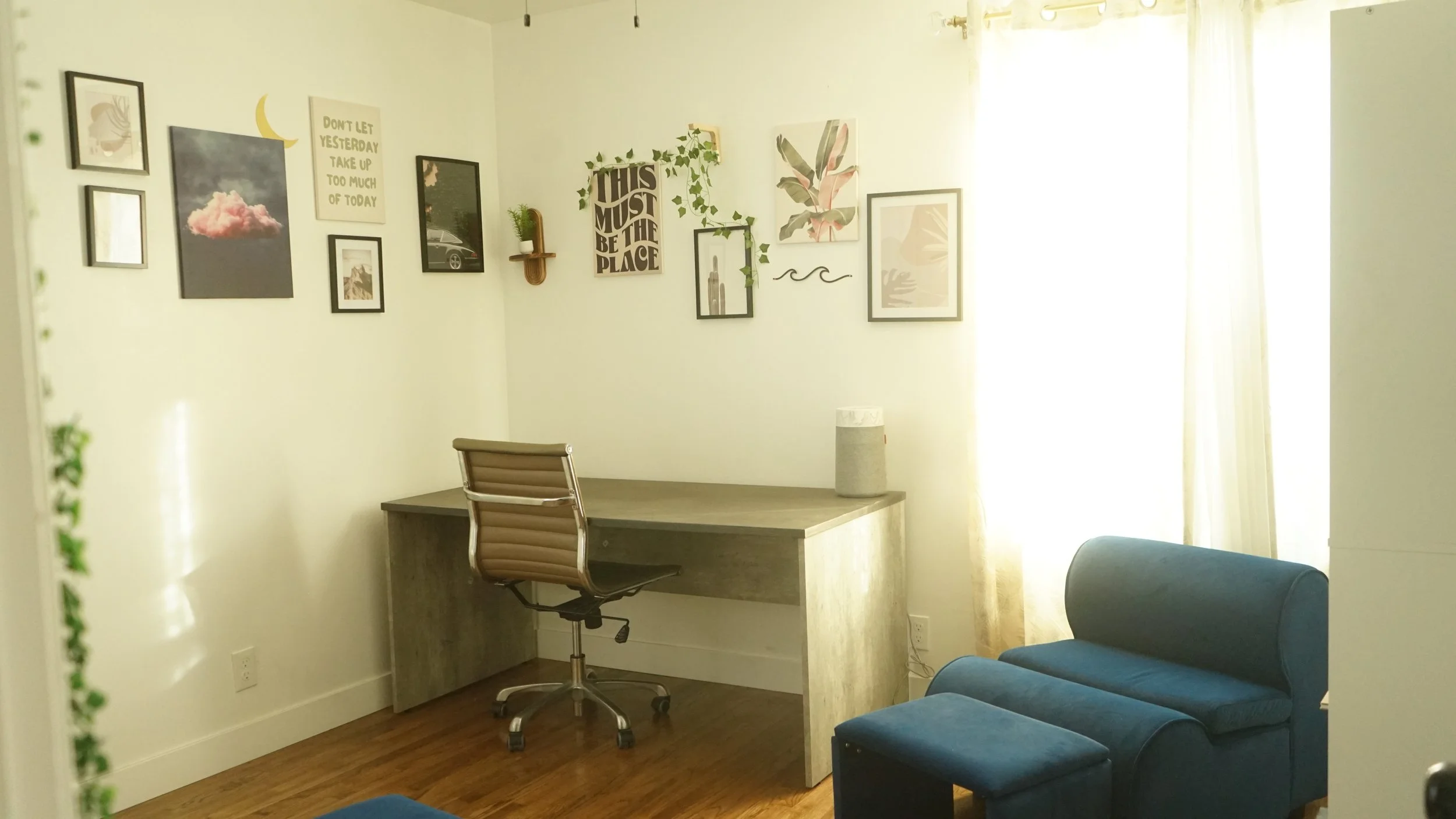 A home office corner with a wooden desk, an office chair, a blue armchair, and a gallery wall with framed pictures and decorative art, illuminated by sunlight from sheer curtains.