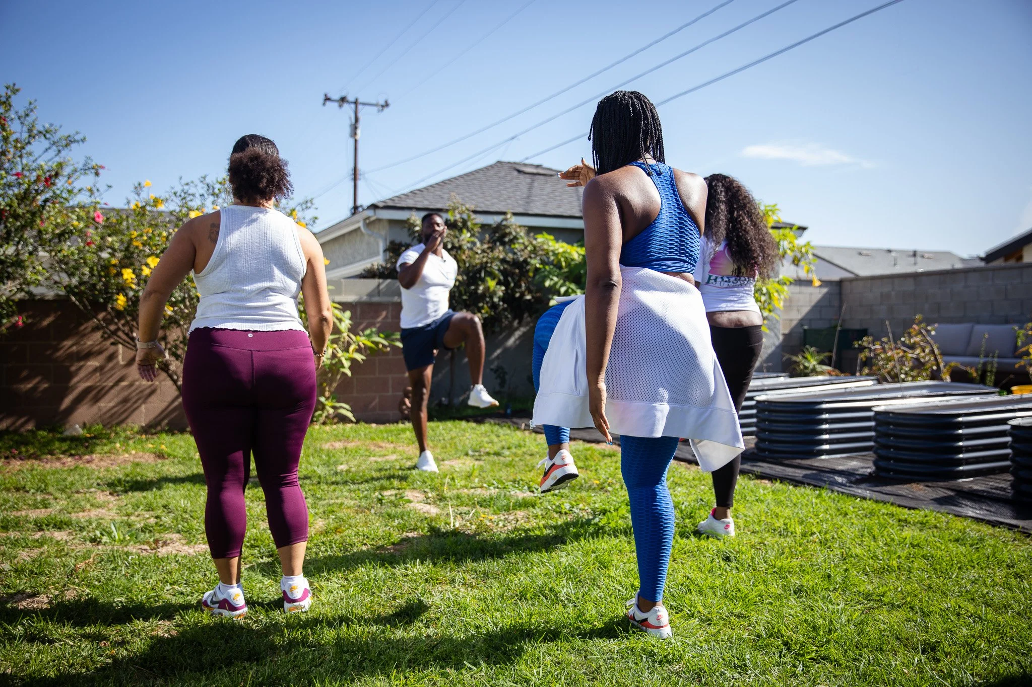 A group of people exercising outdoors in a backyard on a sunny day. They are performing a fitness activity, with some lifting knees and others marching in place, under the supervision of an instructor.
