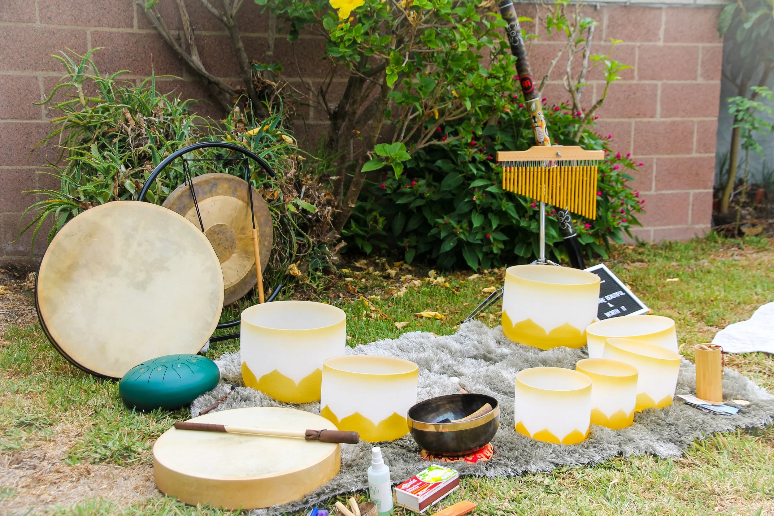 A collection of musical instruments, including tambourines, singing bowls, and a wind chime, arranged on a gray rug outdoors near a garden with plants and a brick wall.