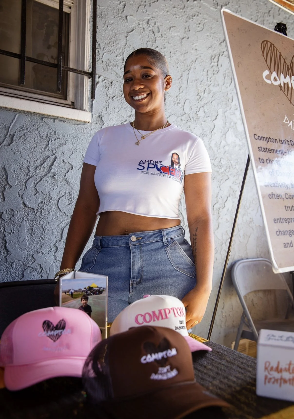 A young woman smiling at a booth with promotional hats and a photo on display, wearing a white crop top and jeans, standing next to a sign with text.