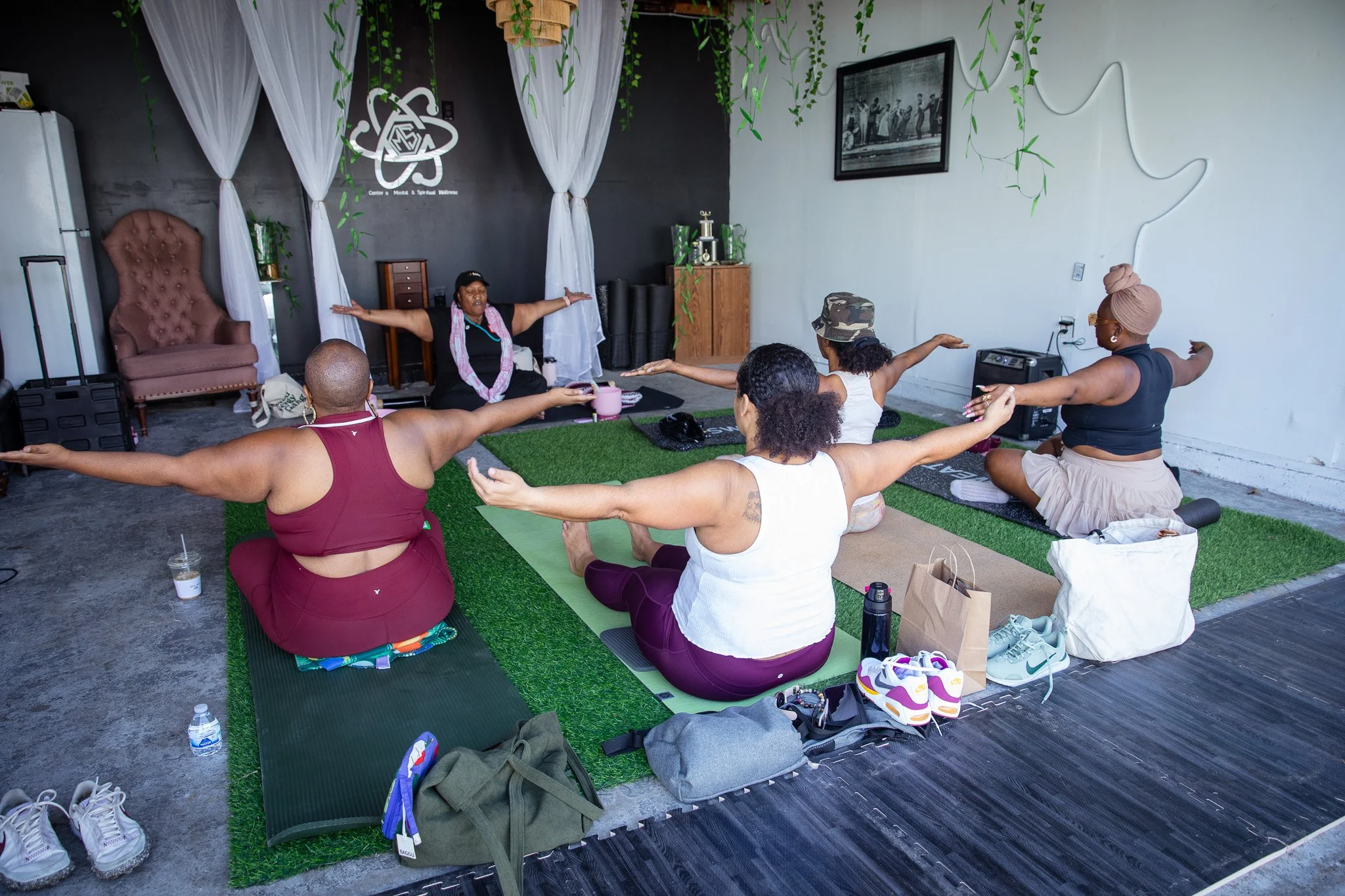 Group of women participating in a yoga class, sitting on mats with arms extended. A instructor leads the session in a room decorated with hanging plants and curtains.