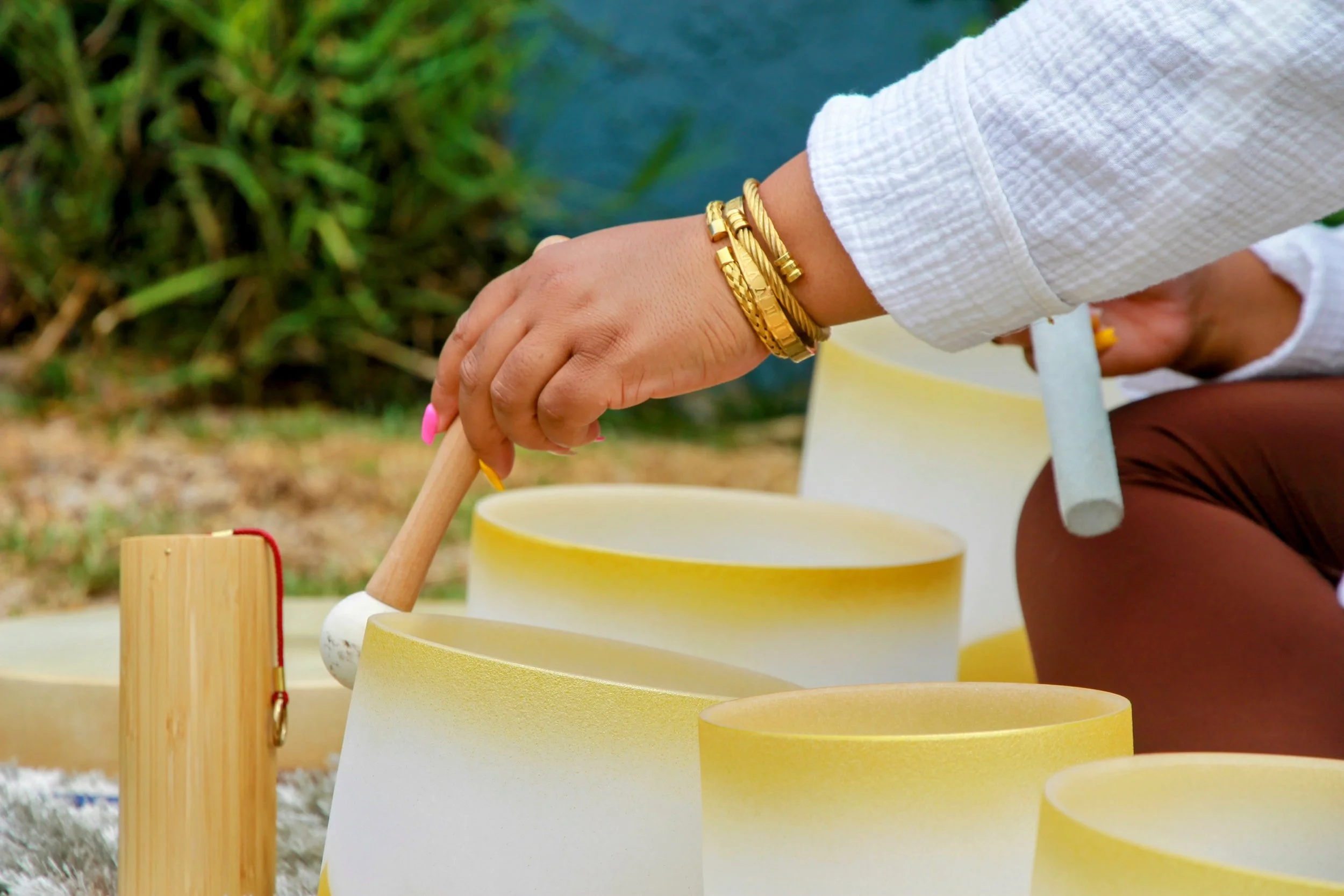 A person wearing gold bracelets plays a xylophone outdoors, with green foliage and a body of water in the background.
