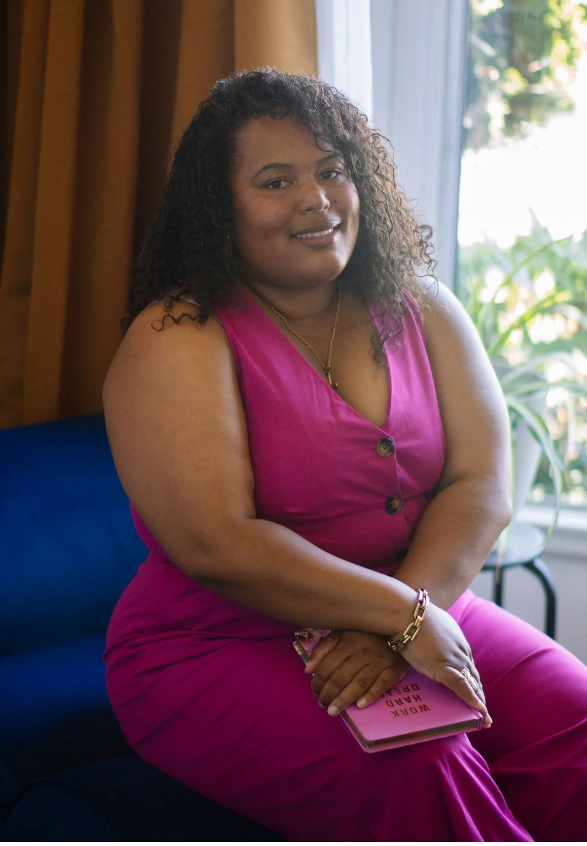 A woman with curly hair sitting on a blue couch, wearing a pink sleeveless dress, holding a small pink notebook titled "Work Hard Dream," in a room with wooden walls and a large window with sunlight and plants outside.
