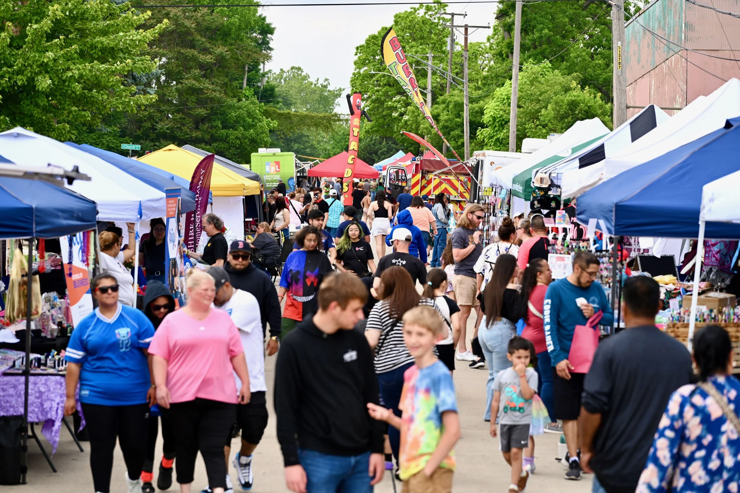 Crowd of people walking through a street fair with vendor tents, green trees, and a fire truck in the background.