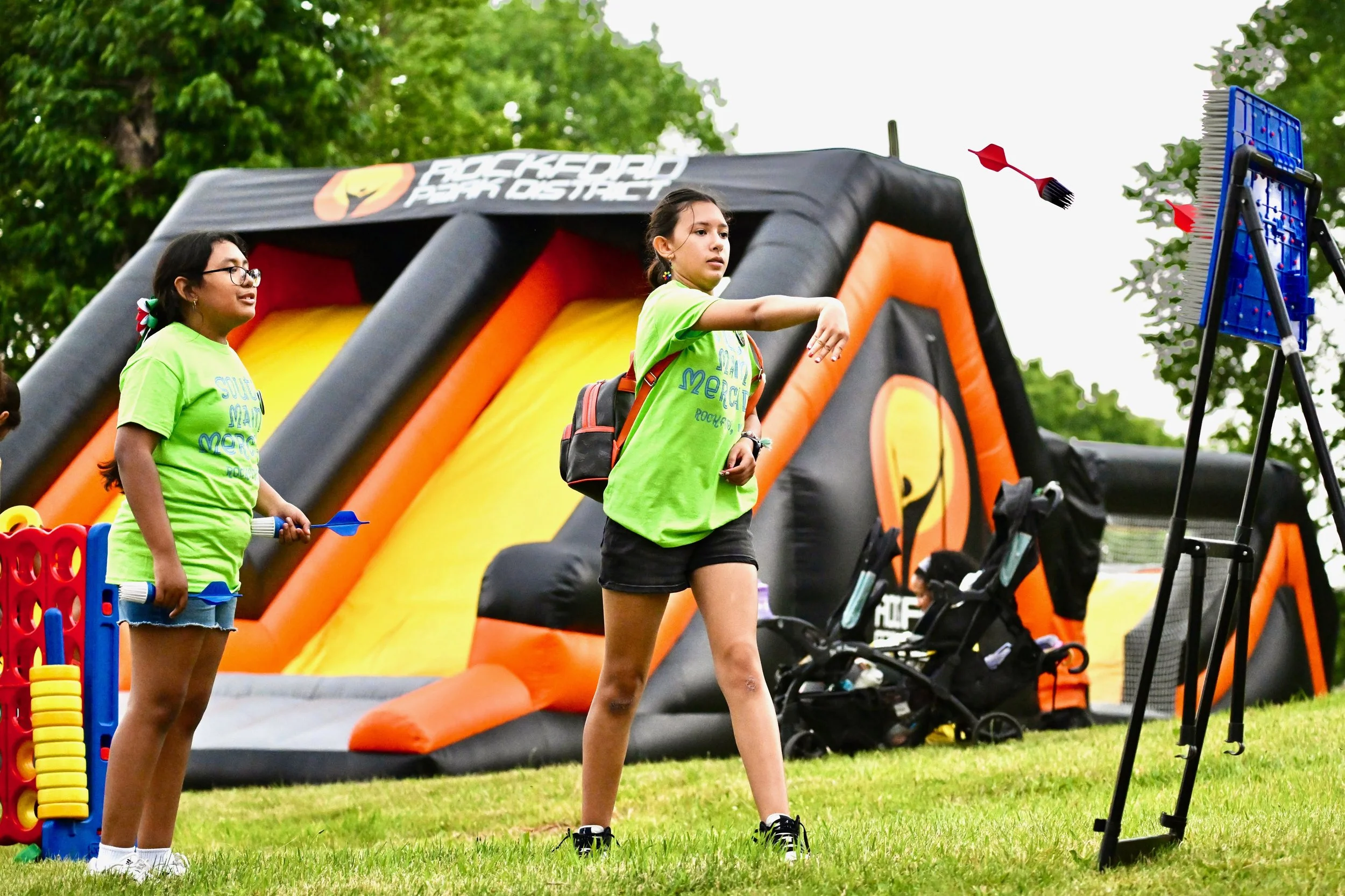 Two young girls participating in a dart-throwing game at an outdoor carnival or festival, with a large inflatable climbing wall and trees in the background.