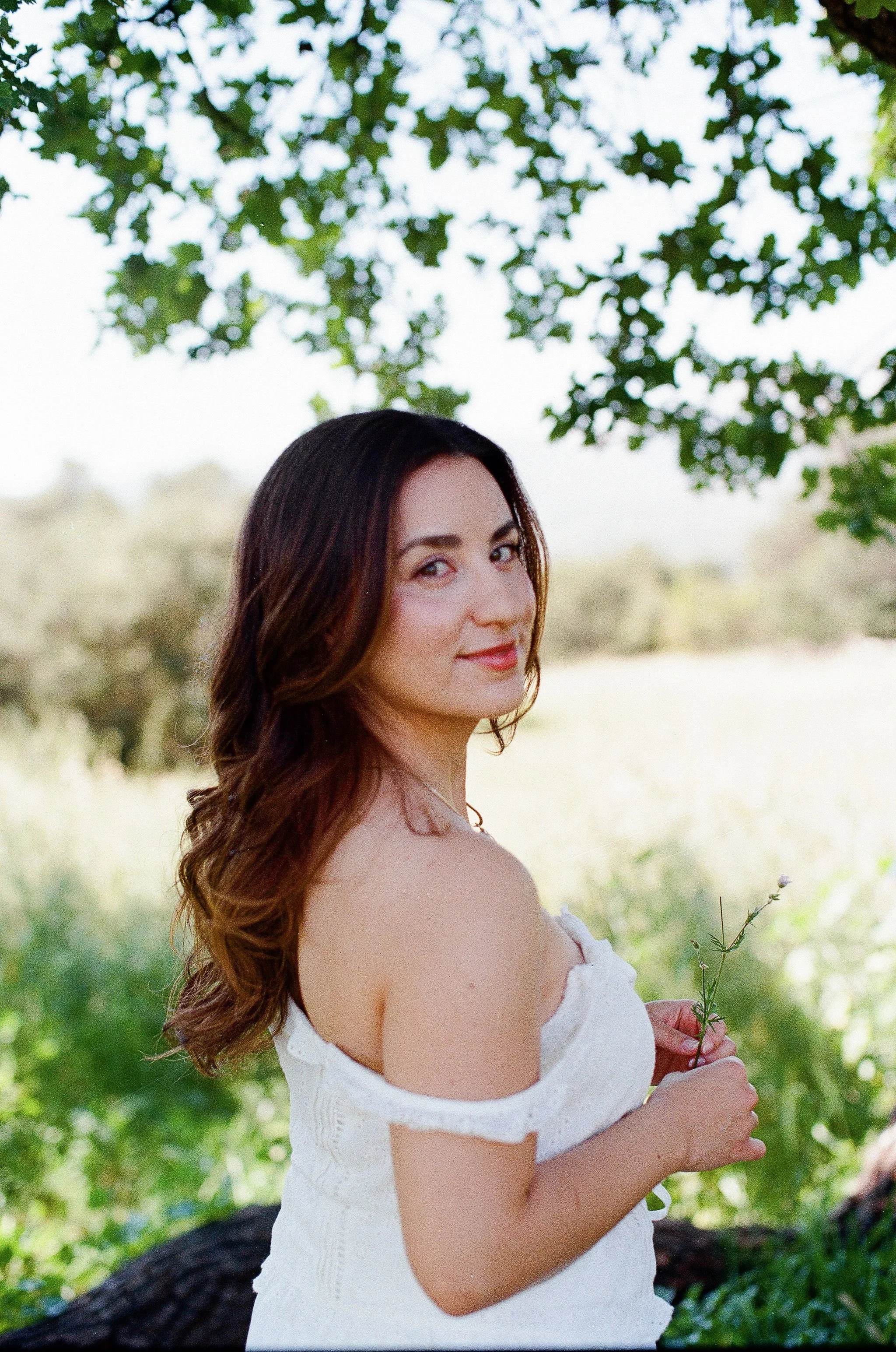 A woman with long brown hair and fair skin wearing a white off-the-shoulder dress standing outdoors under green trees, holding a small flower.
