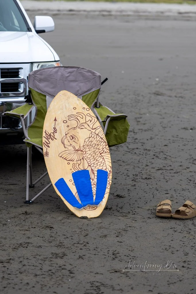image of a laser engraved wooden skimboard propped against a folding chair and some sandals in the sand. Grayland, Washington