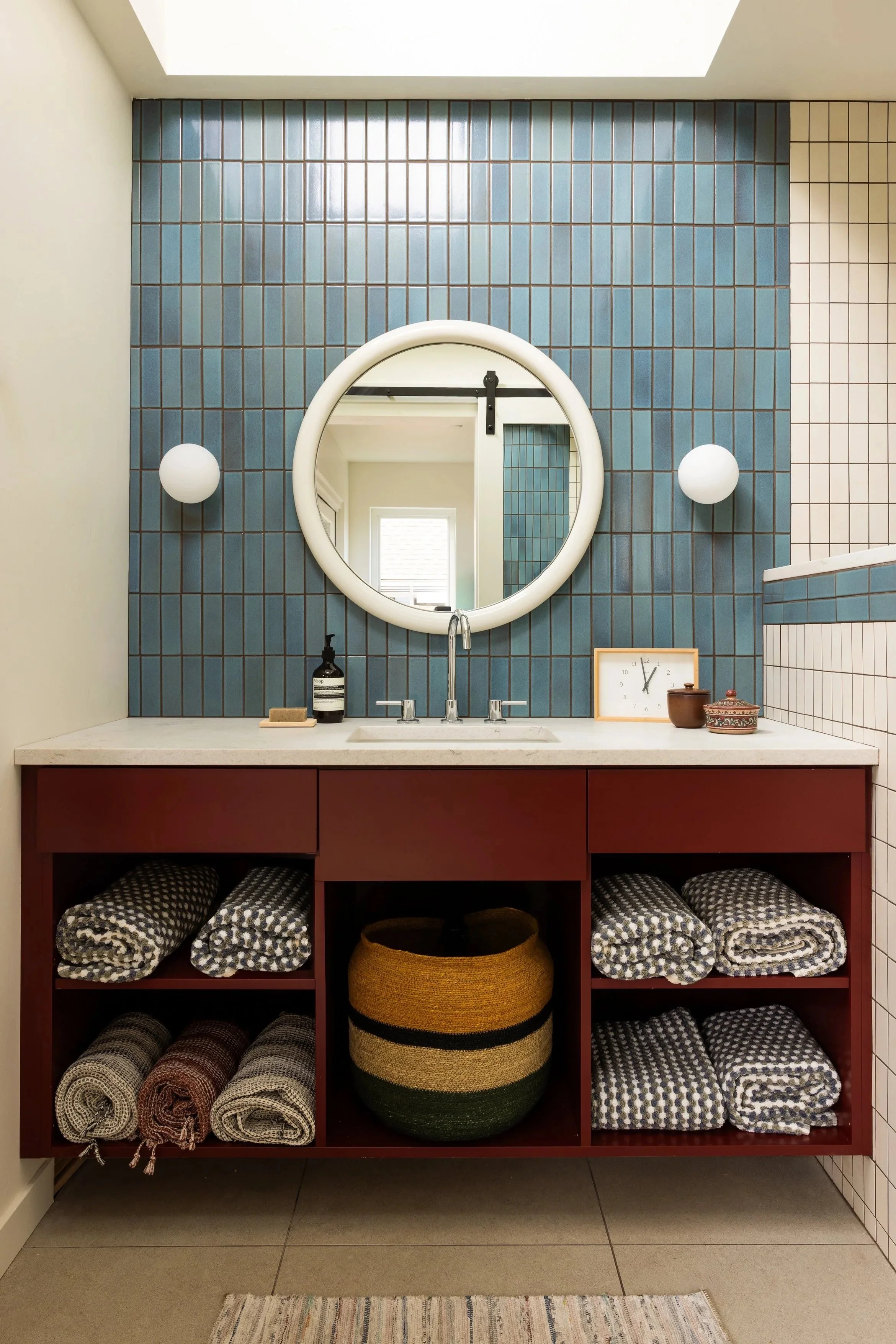 Bathroom with blue tiled accent wall, white round mirror, black and white soap dispenser, clock, and towels stored in open shelves on a red cabinet under the sink.