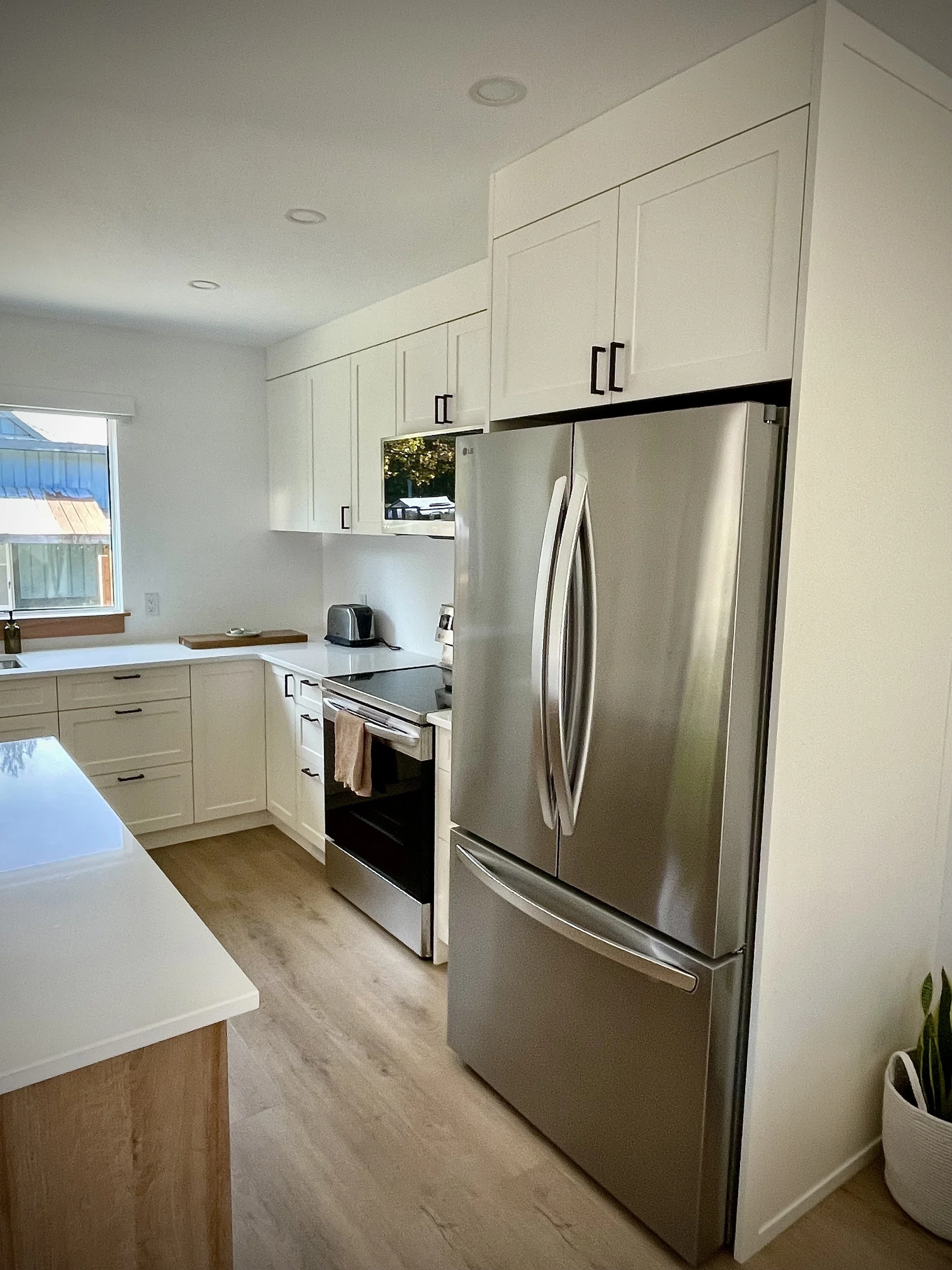 Modern kitchen with white cabinets, stainless steel refrigerator, stove, toaster, and wood flooring, near a window with a view outside.