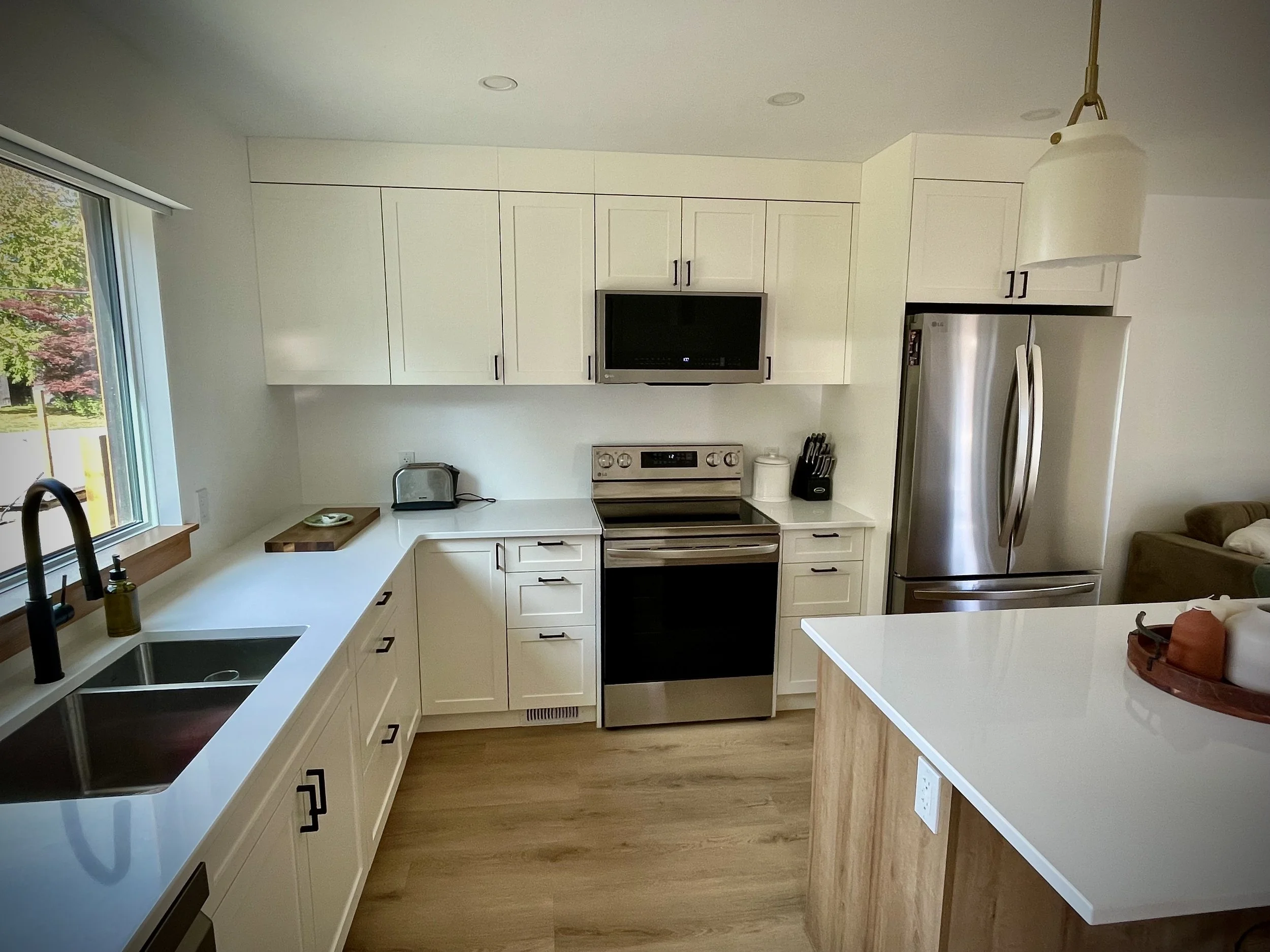 Modern kitchen with white cabinets, stainless steel refrigerator and oven, microwave, black faucet, window with view of trees, white countertop, toaster, knife block, and decorative tray with vases.