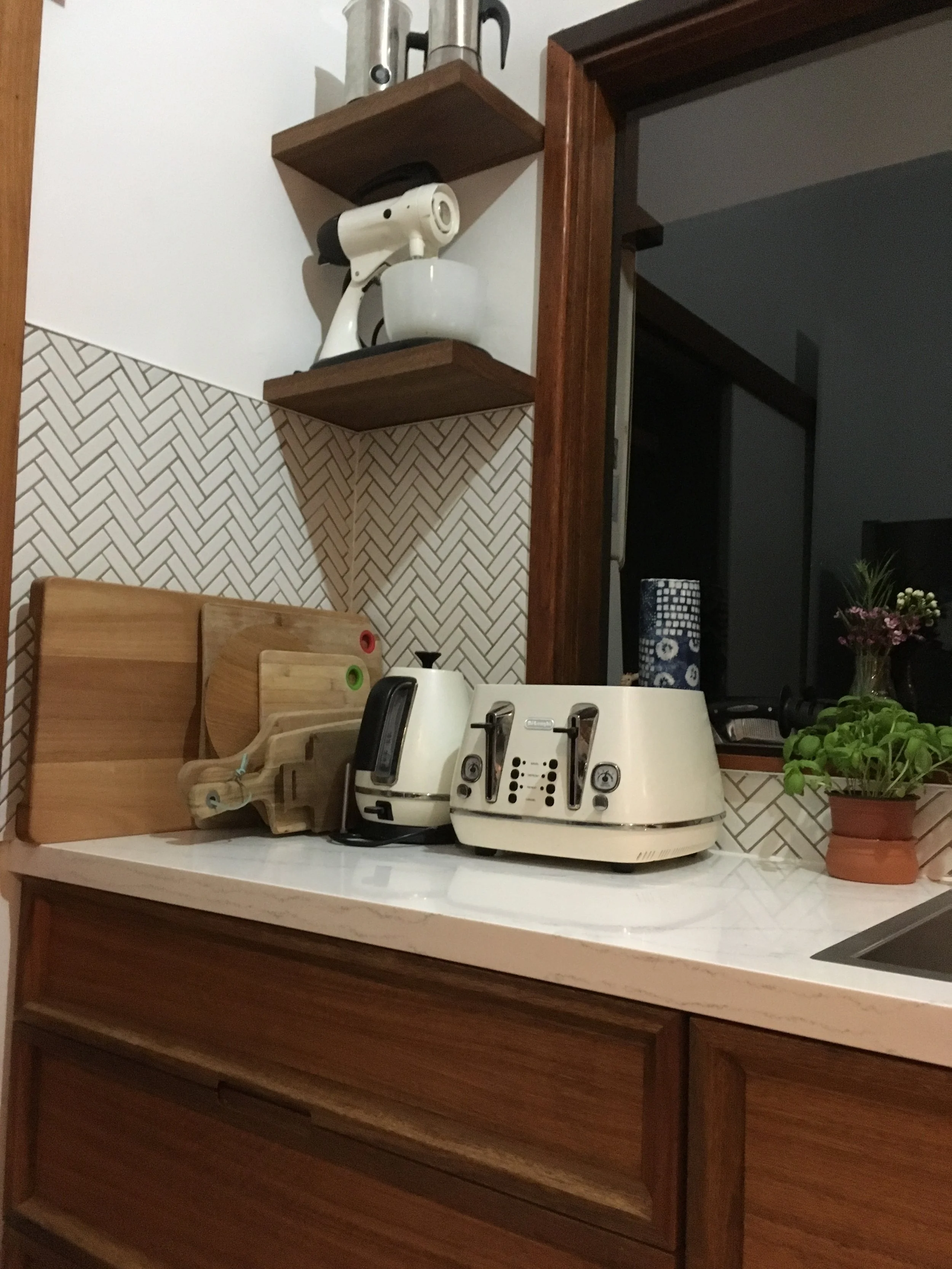 Kitchen countertop with vintage beige two-slice toaster, a black landline phone, and a small potted basil plant, with wooden cutting boards and a white coffee grinder nearby, and a cherry red knife on a wooden wall-mounted rack, in a kitchen with bro