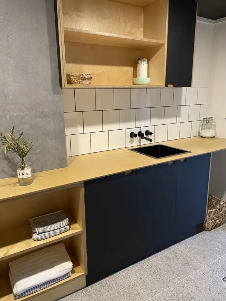 A minimalist kitchen with a wooden countertop, open shelves, white tile backsplash, black faucet and sink, and closed black and wood cabinets. Decor includes a glass jar, a small plant, and folded towels.