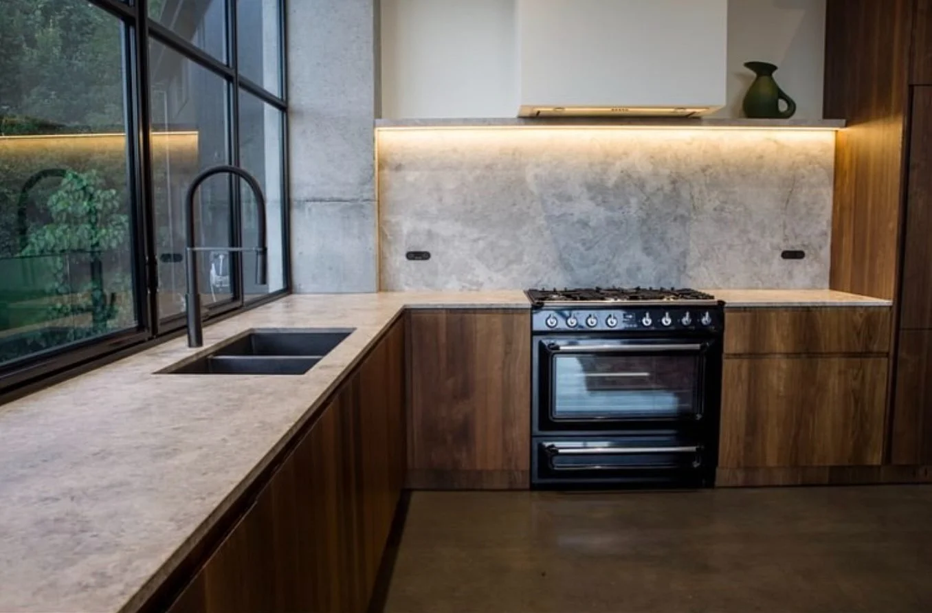 Modern kitchen with wood cabinets, a black stove, marble countertops, a subway tile backsplash, a black sink, and large glass windows with a view of greenery outside.