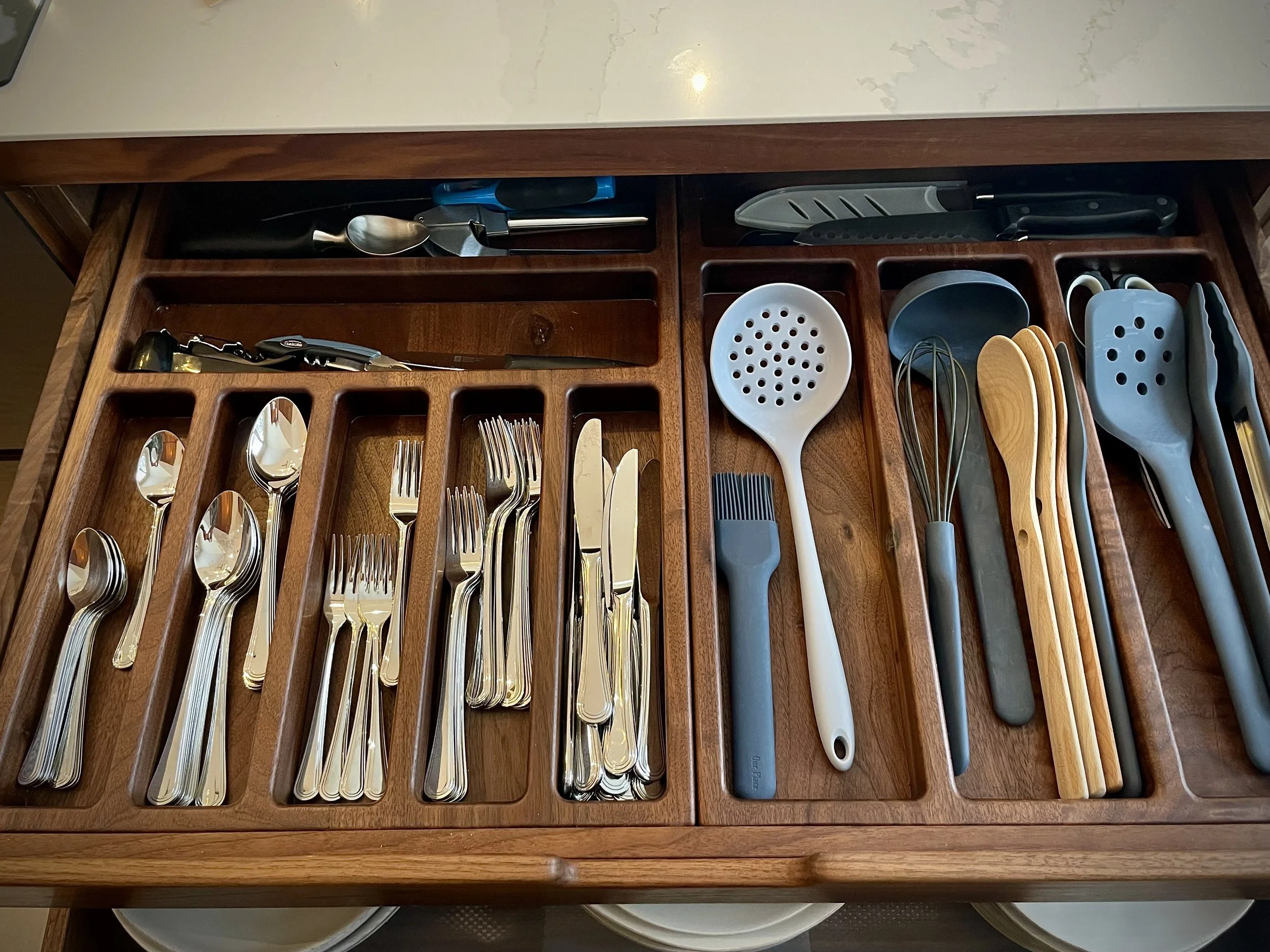 Drawer filled with assorted kitchen utensils including spoons, forks, knives, a slotted spoon, whisk, spatulas, tongs, and various other cooking tools.