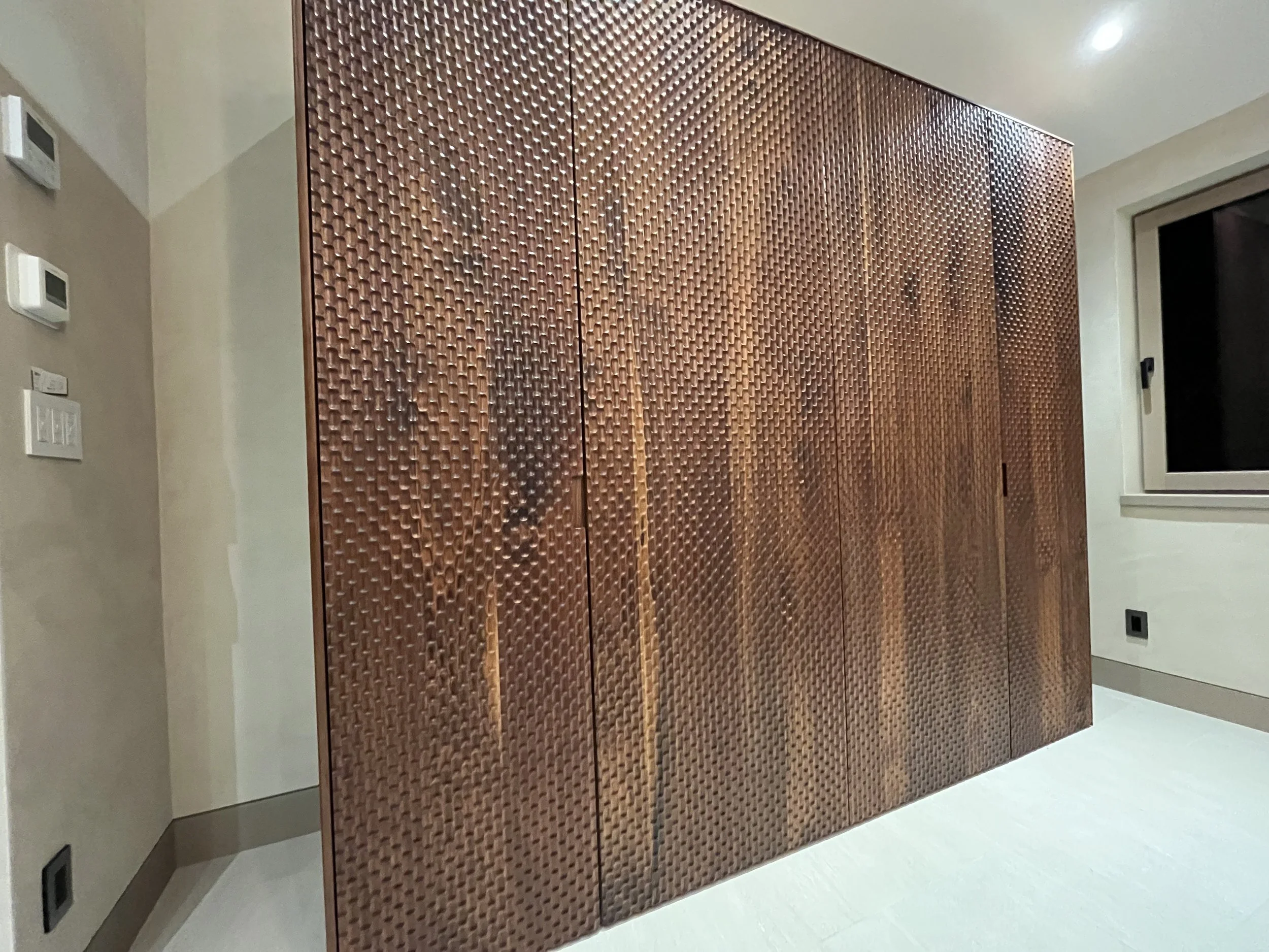 A large, textured, copper-colored wardrobe with a repeated geometric pattern in a room with beige walls, a window, and a thermostat.