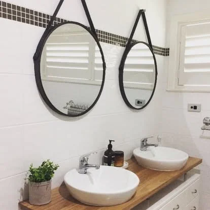 Two round mirrors with black frames are mounted on a white tiled bathroom wall above a wooden countertop with two white vessel sinks. A plant is placed on the left side of the countertop.