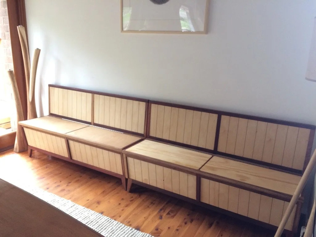 Unfinished wooden bench or storage unit with multiple compartments, placed against a white wall in a room with wooden floors and curtains.