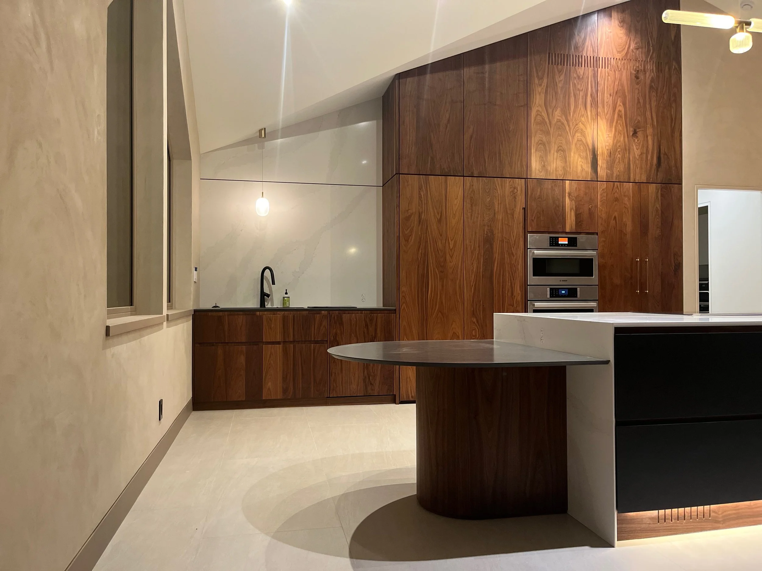Modern kitchen with beige walls, light flooring, and dark wood cabinets. There is a peninsula countertop with dark and white surfaces and a built-in oven, microwave, and oven stacked in the cabinetry. A small window with beige trim and a single pendant light hanging from the ceiling are also visible.