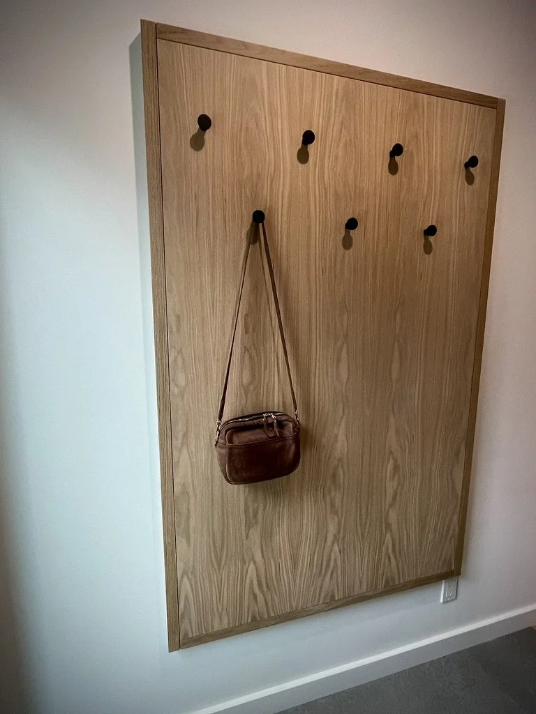 Wooden wall panel with six black hooks, one hook holding a brown shoulder bag, against a white wall.
