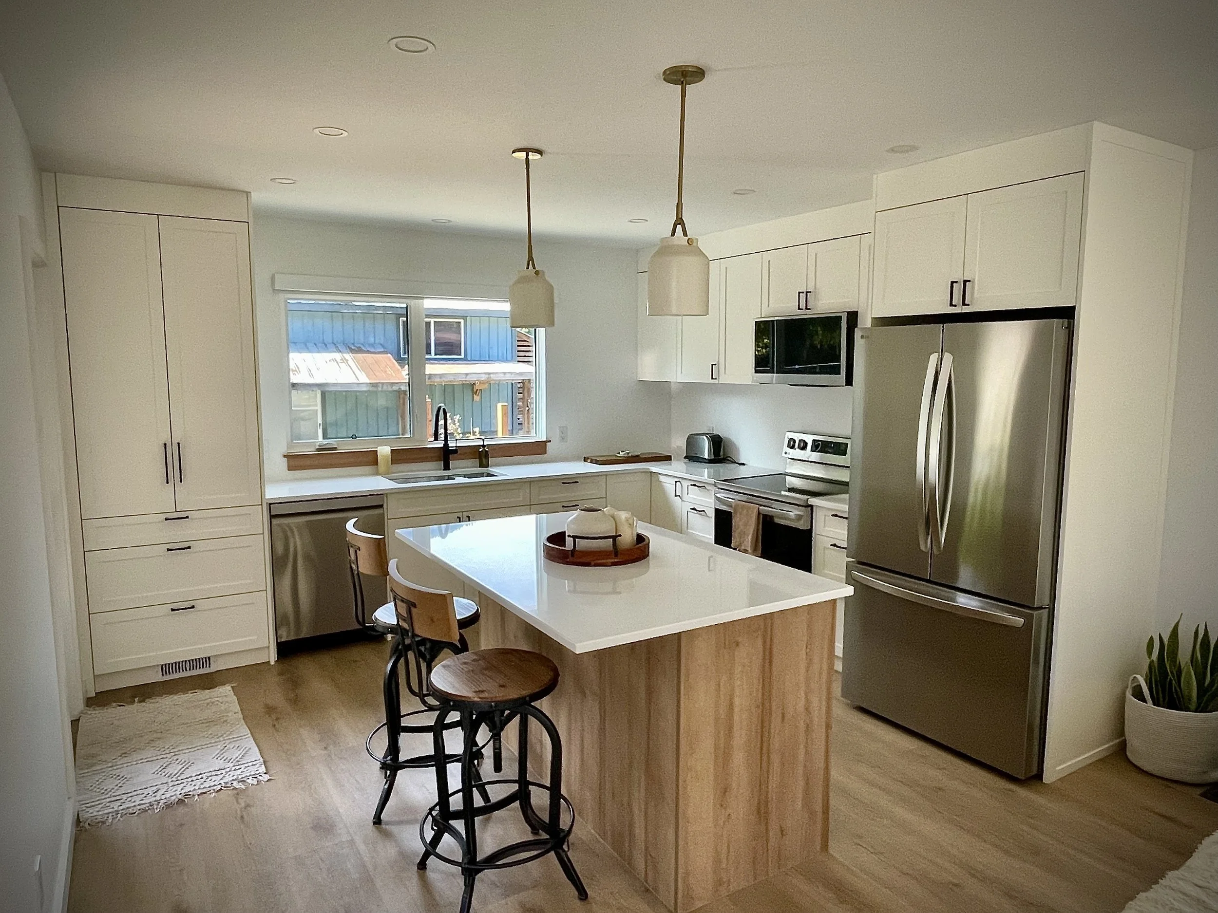 Modern kitchen with white cabinets, stainless steel refrigerator, island with wooden base, two hanging pendant lights, and a window overlooking neighboring houses.