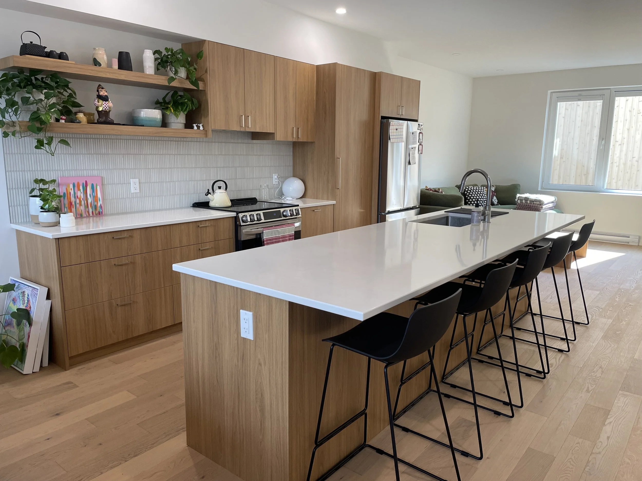 Modern kitchen with wooden cabinets, white countertops, a large island with seating, a stainless steel refrigerator, and a window letting in natural light.