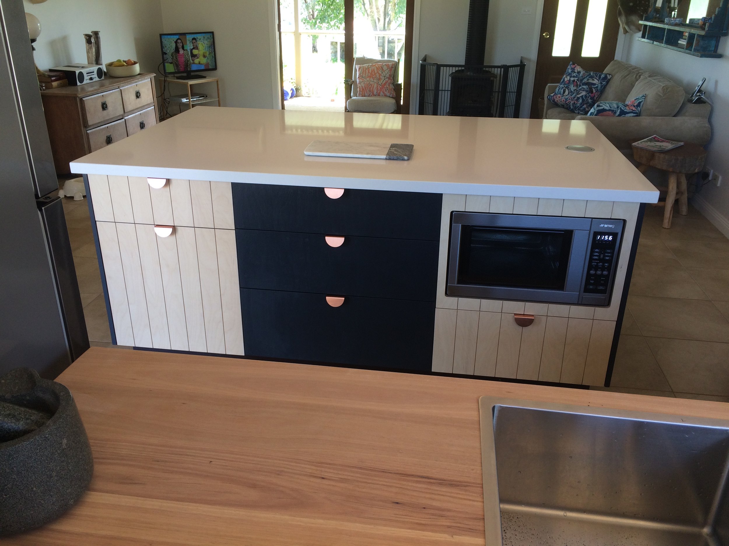 A kitchen island with a white countertop, with a black and natural wood-paneled cabinet below. A microwave is built into the right side of the island. In the background, there is a living room with a sofa, a chair, a small table with a book, and a wo