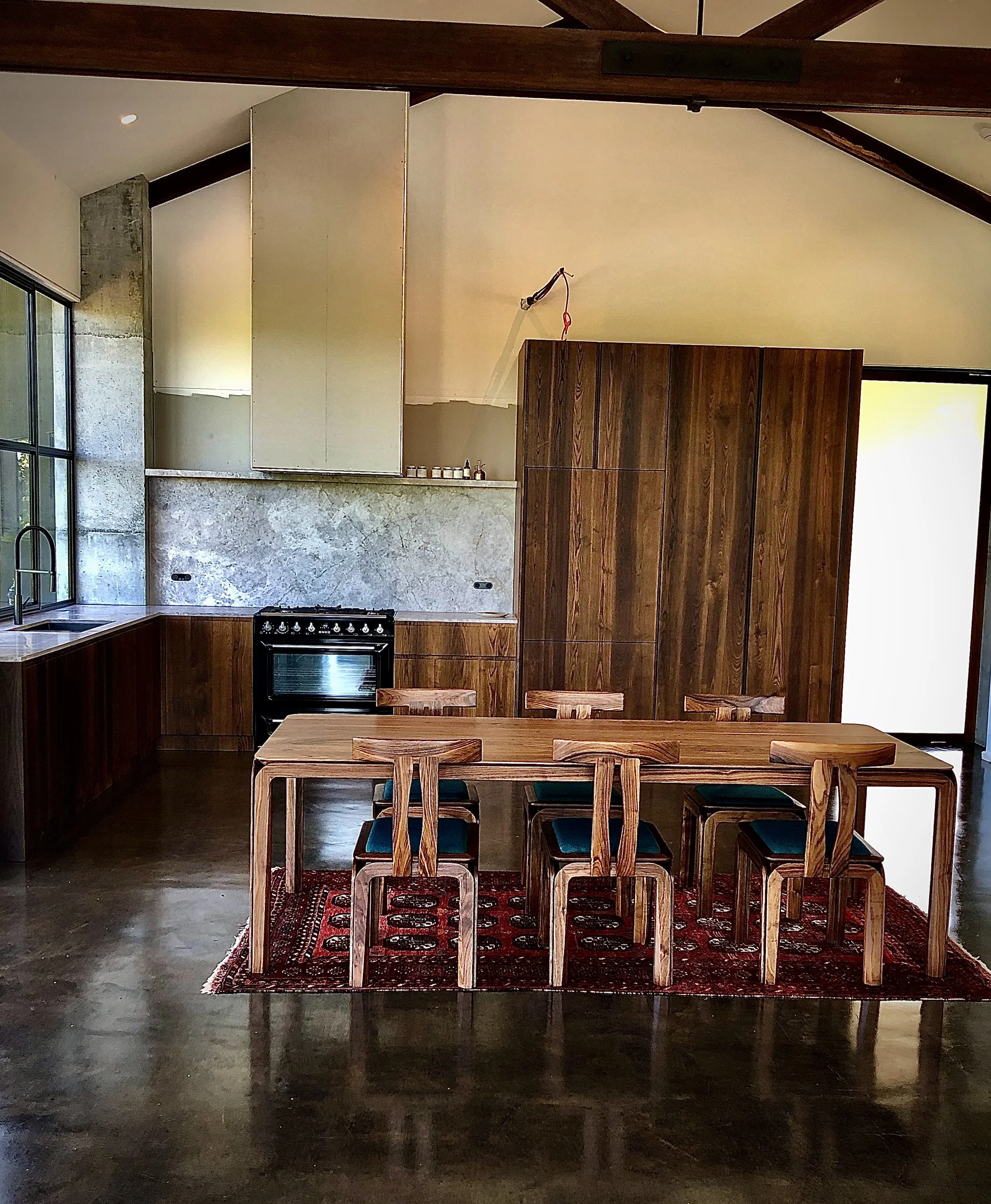 Modern kitchen with wooden cabinets, a dining table with six chairs, and a red patterned rug. The kitchen features a black oven, a marble backsplash, and large windows.