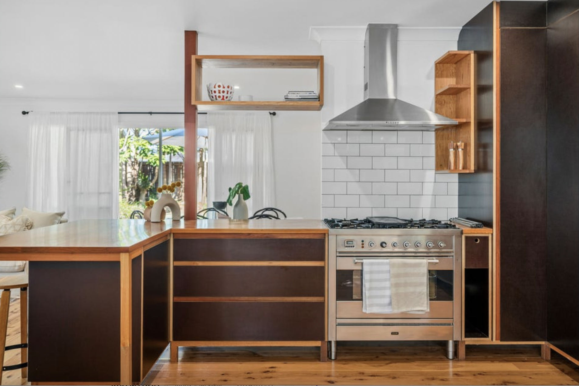 Modern kitchen with wooden accents, stainless steel stove and hood, white subway tile backsplash, open shelving, and a view of a bright dining area with large windows and white curtains.