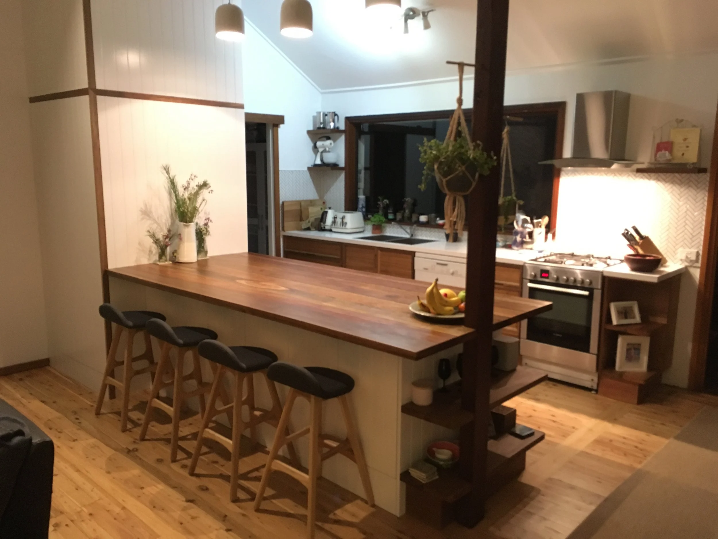 A cozy kitchen with a large wooden island countertop, four bar stools, a vase with flowers, banana and fruit bowl, and various appliances and decor.