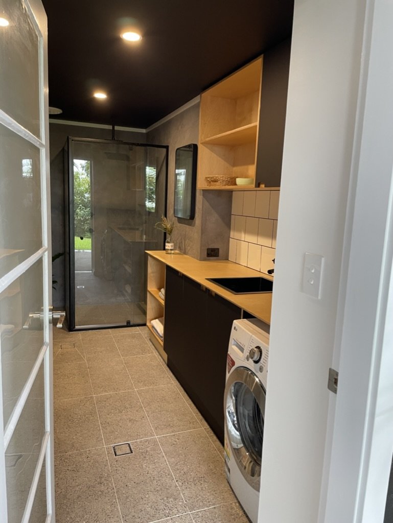 A modern laundry room with beige tiles, a wooden countertop, open shelves, a washing machine, and a glass-enclosed shower at the end.