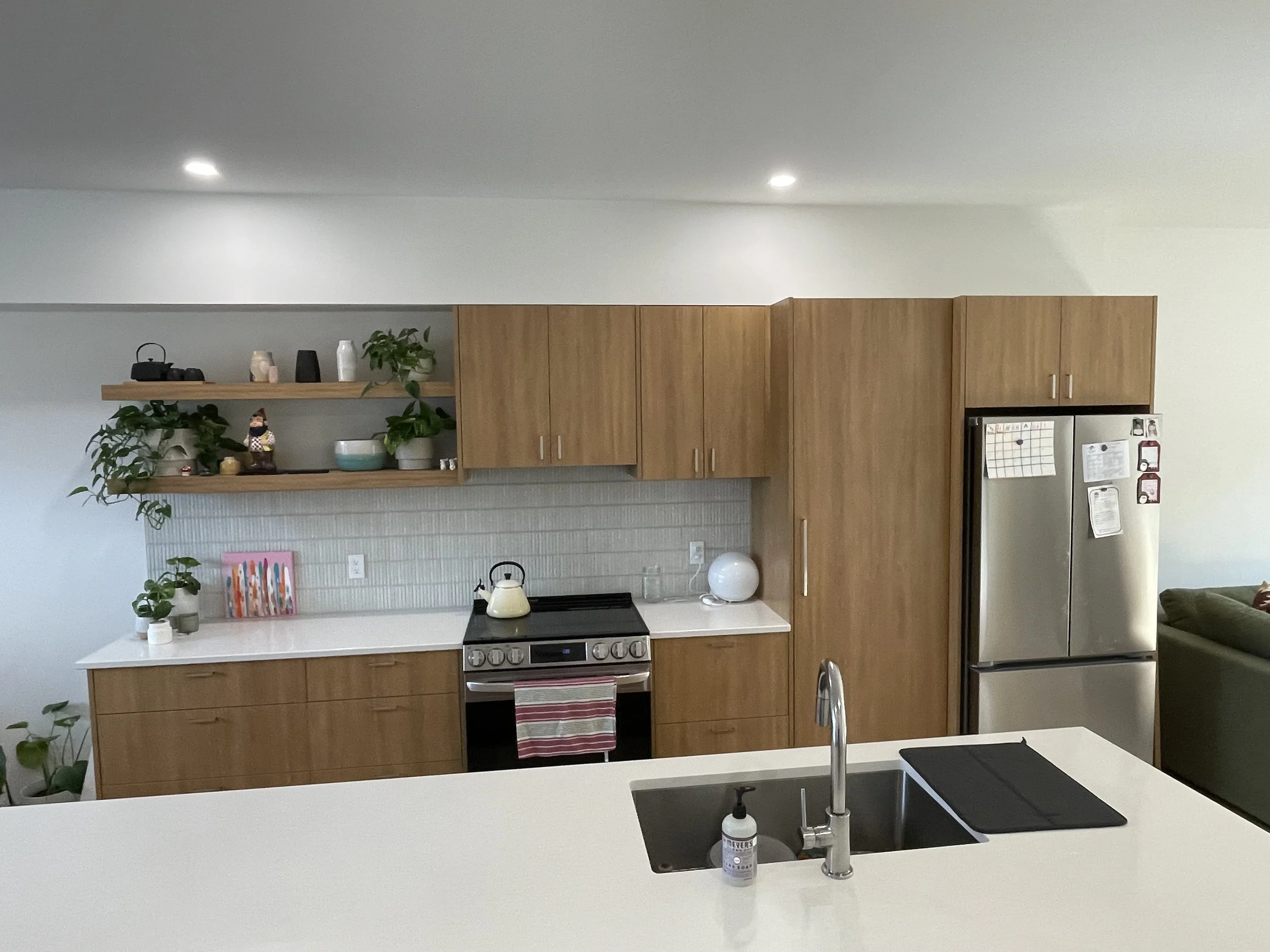 Modern kitchen with white countertops, wooden cabinets, stainless steel refrigerator, black oven, and green plants on open shelves.