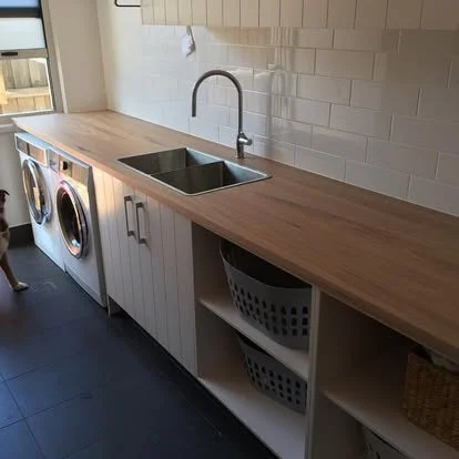 A laundry room with a wooden countertop, a double sink, a faucet, a washing machine and a dryer, open shelves under the counter, and a white wall with tiles.