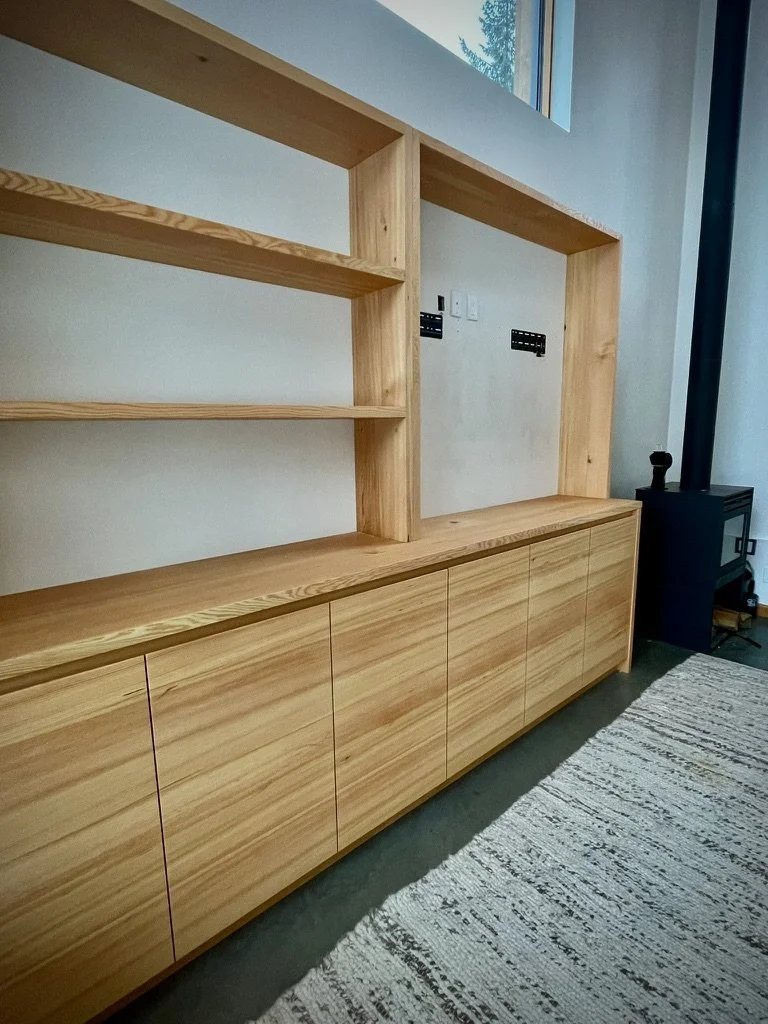 A modern wooden built-in cabinet with open shelving and closed storage at the bottom, situated against a wall near a window with a view of trees.