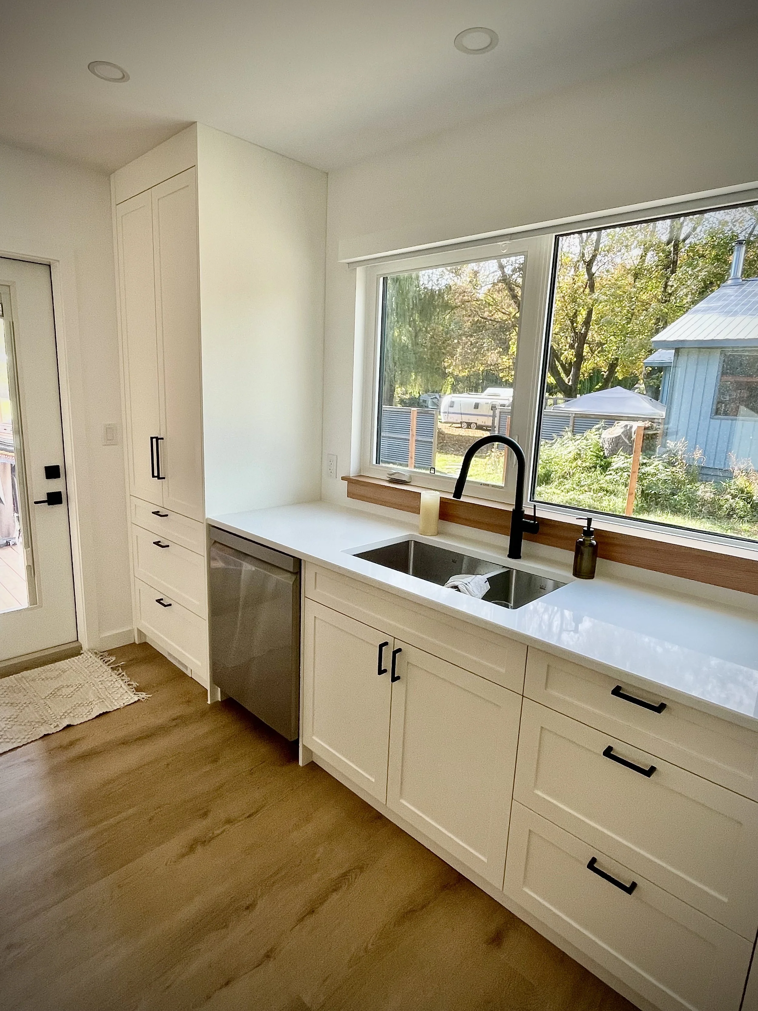 Modern kitchen with white cabinets, a large window above the sink showing an outdoor view, and hardwood floors.