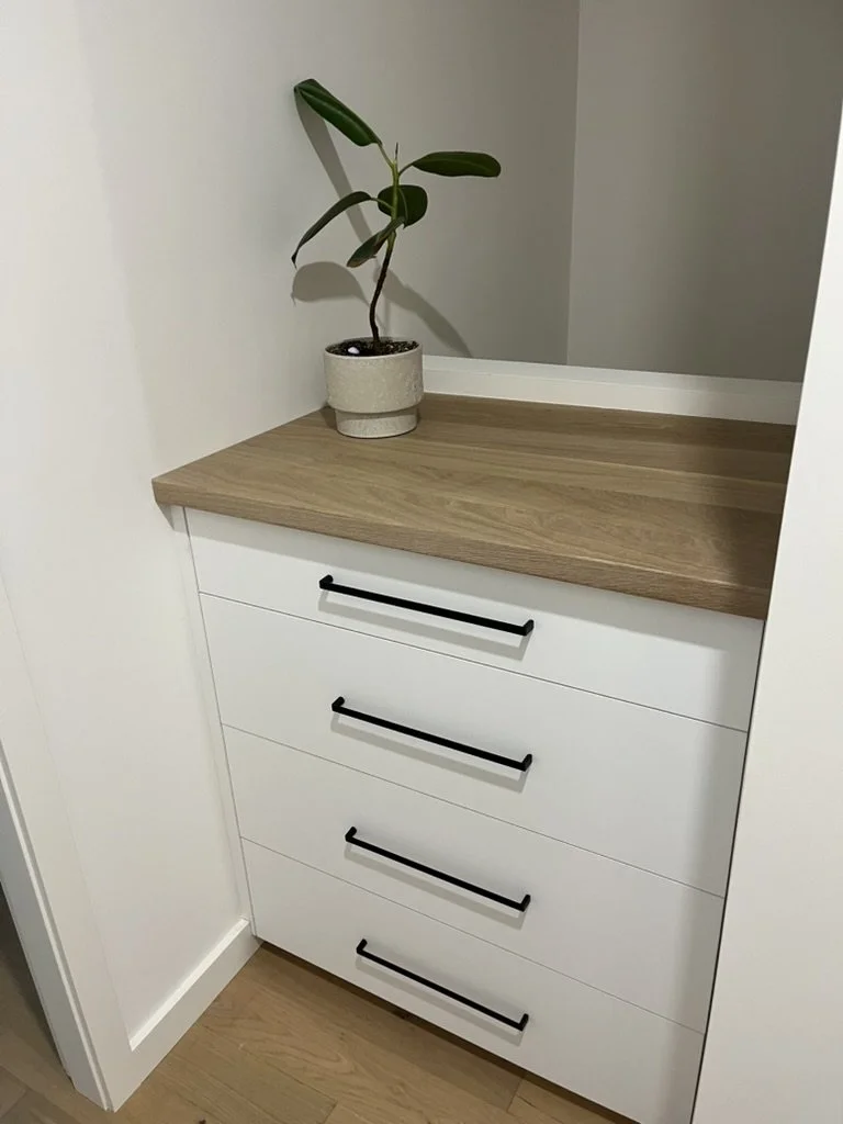 A white dresser with four drawers and black handles, topped with a light wood surface. A potted plant with broad green leaves sits on top of the dresser in the corner of a room.