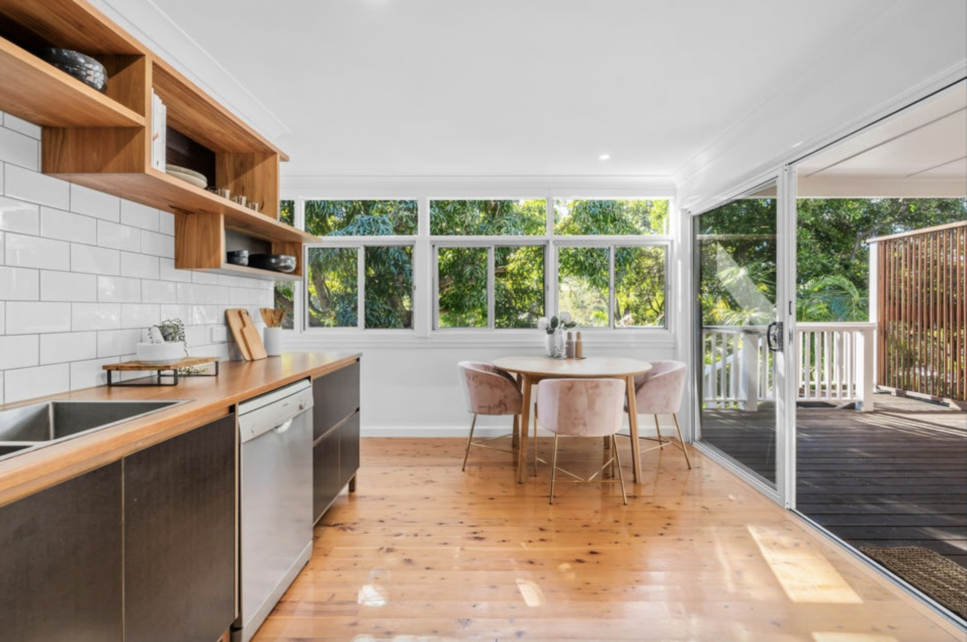 Kitchen with wooden cabinets, a round dining table with pink chairs, large windows with green foliage outside, and a sliding glass door leading to a wooden balcony.