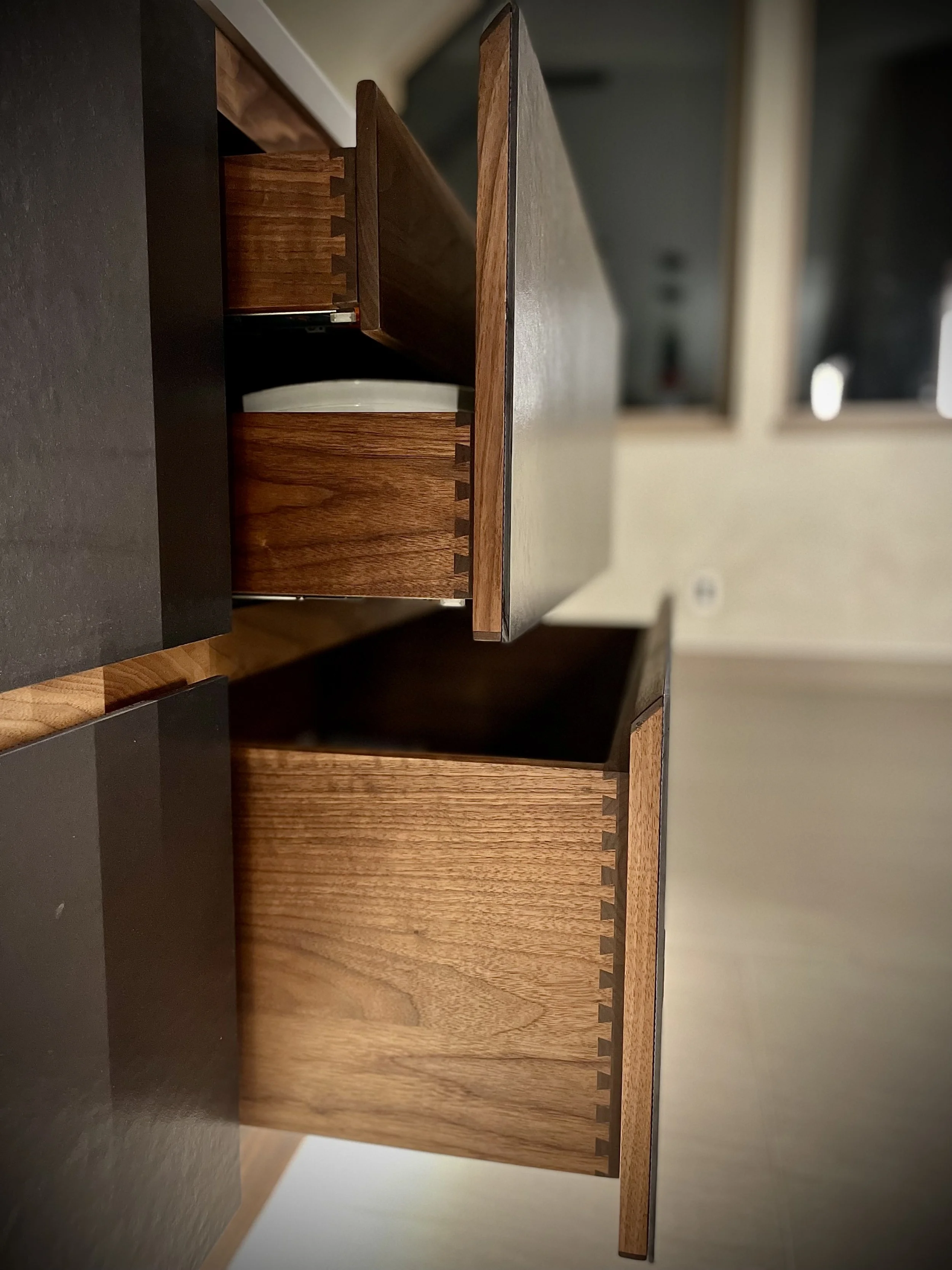 Close-up of an open wooden cabinet drawer with a white plate inside, showing dovetail joints and dark interior, in a modern kitchen.